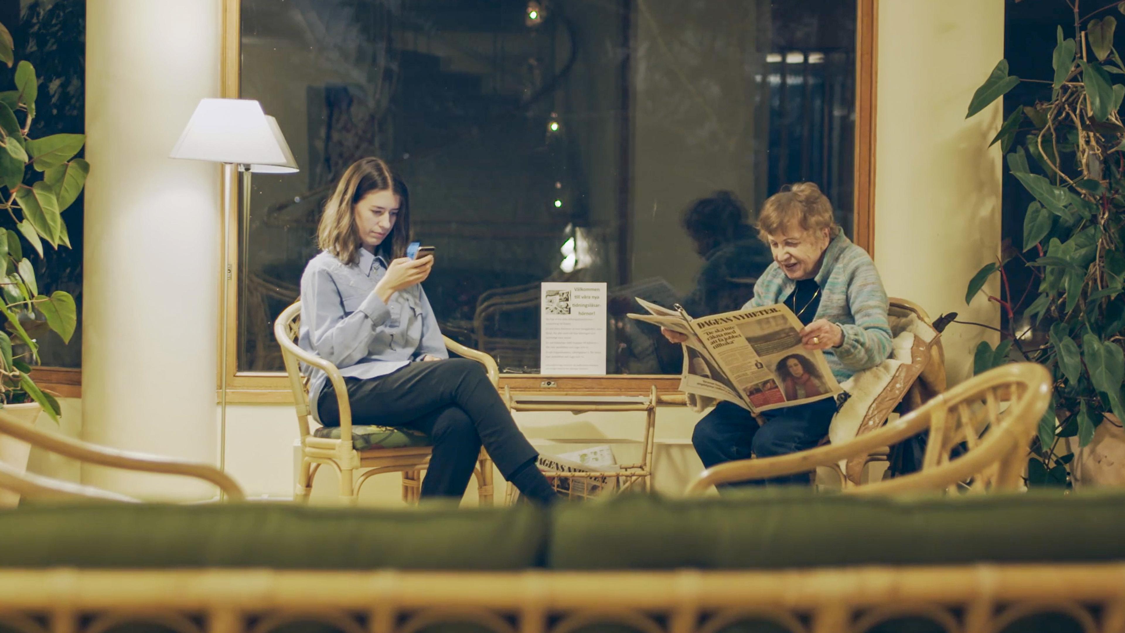 Photo of two women seated indoors, one using a phone the other reading a newspaper, with plants and wooden furniture.