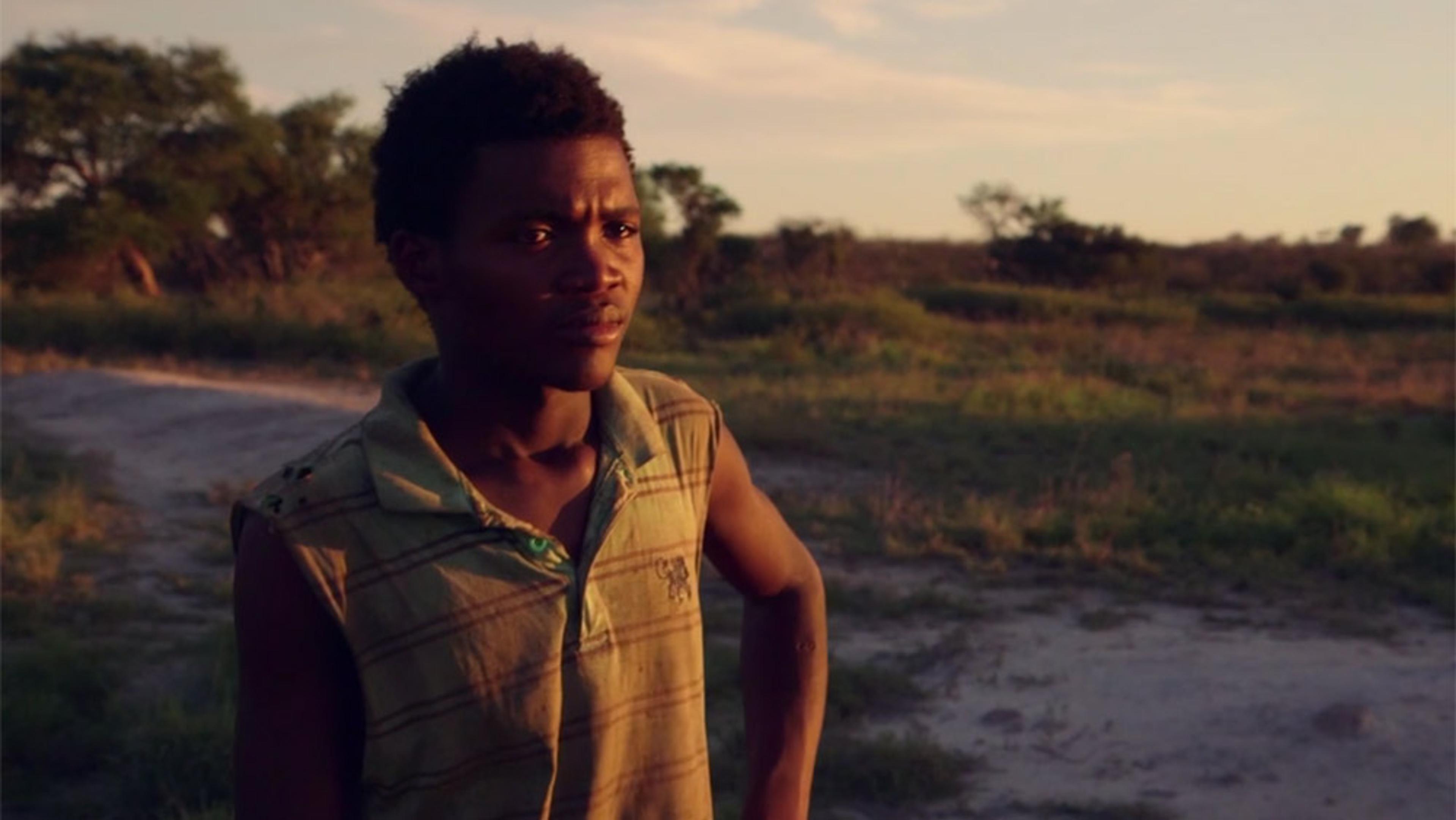A young man in a sleeveless shirt standing in a rural area at sunset, surrounded by trees and grass.