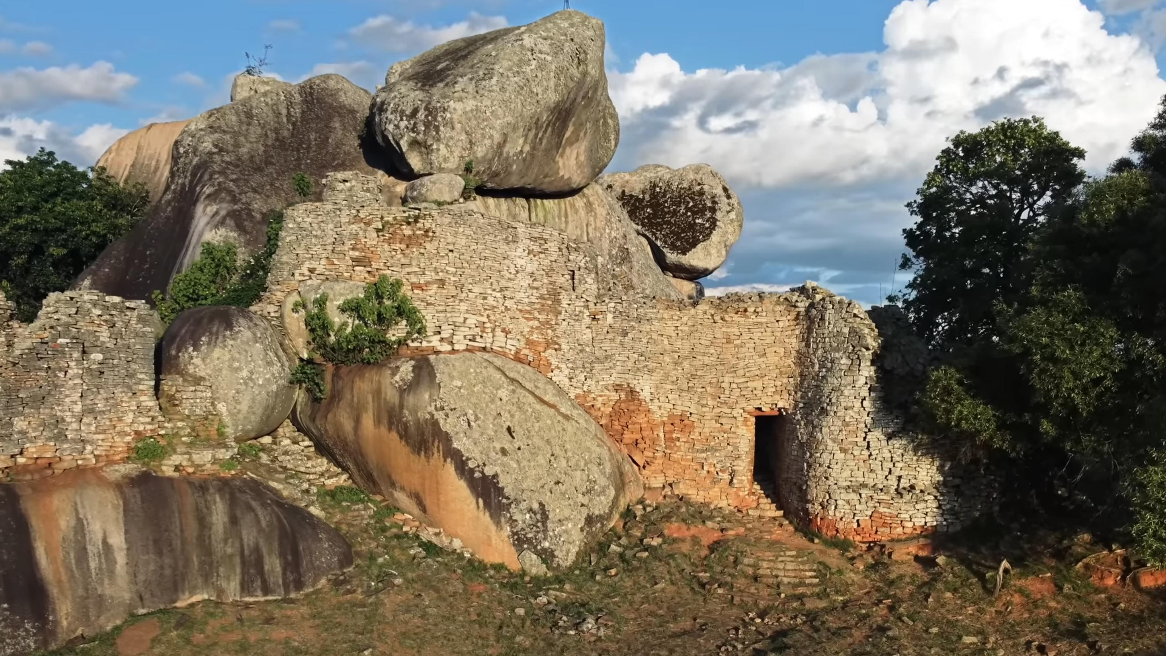 Photo of ancient stone ruins with large boulders on top surrounded by trees under a blue sky with clouds.