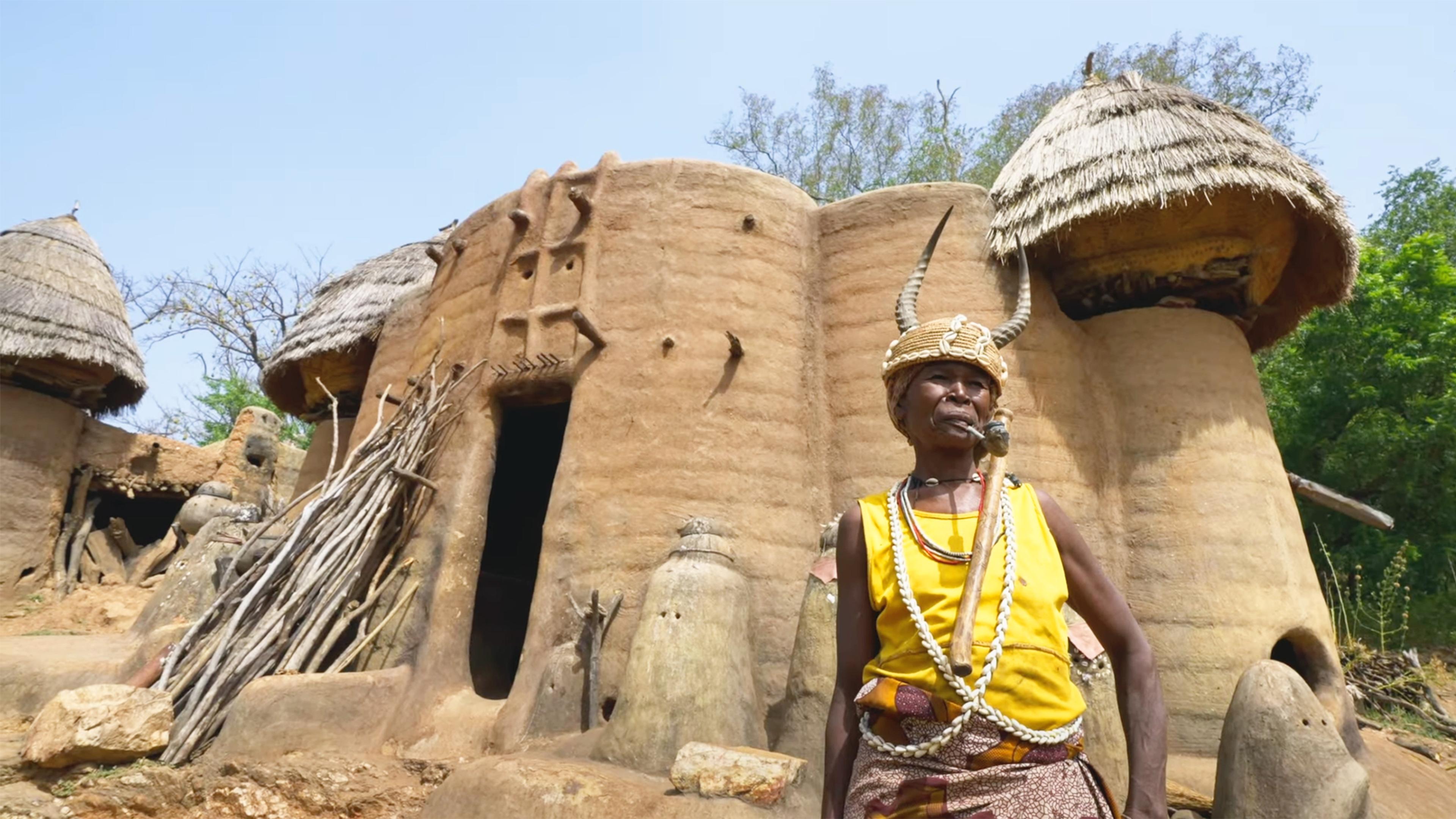 Photo of a person wearing traditional headwear with animal horns standing before clay huts with thatched roofs in a rural setting in Western Africa.