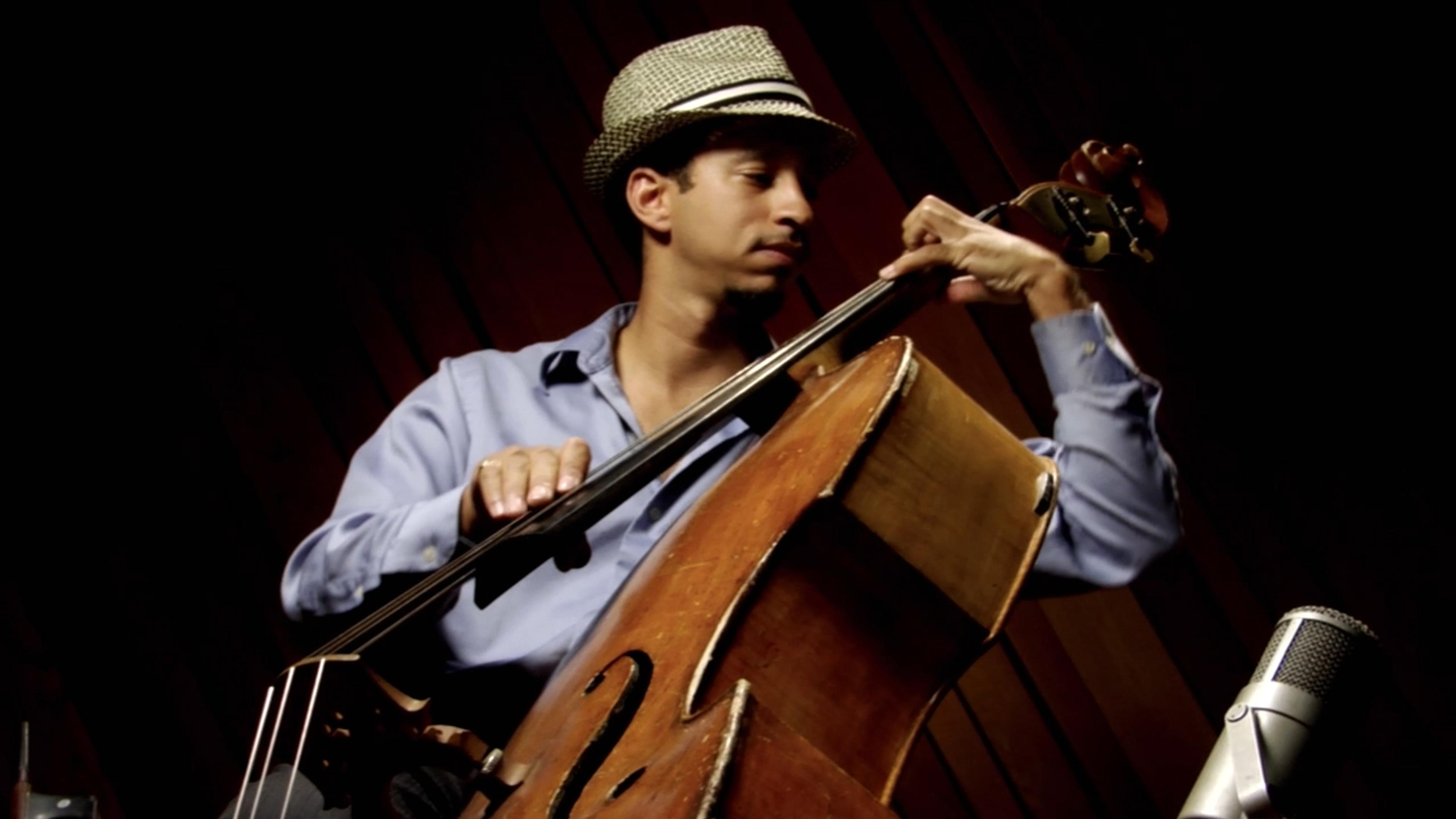 Photo of a musician in a hat playing a double bass with a microphone in front, set against a dark background.