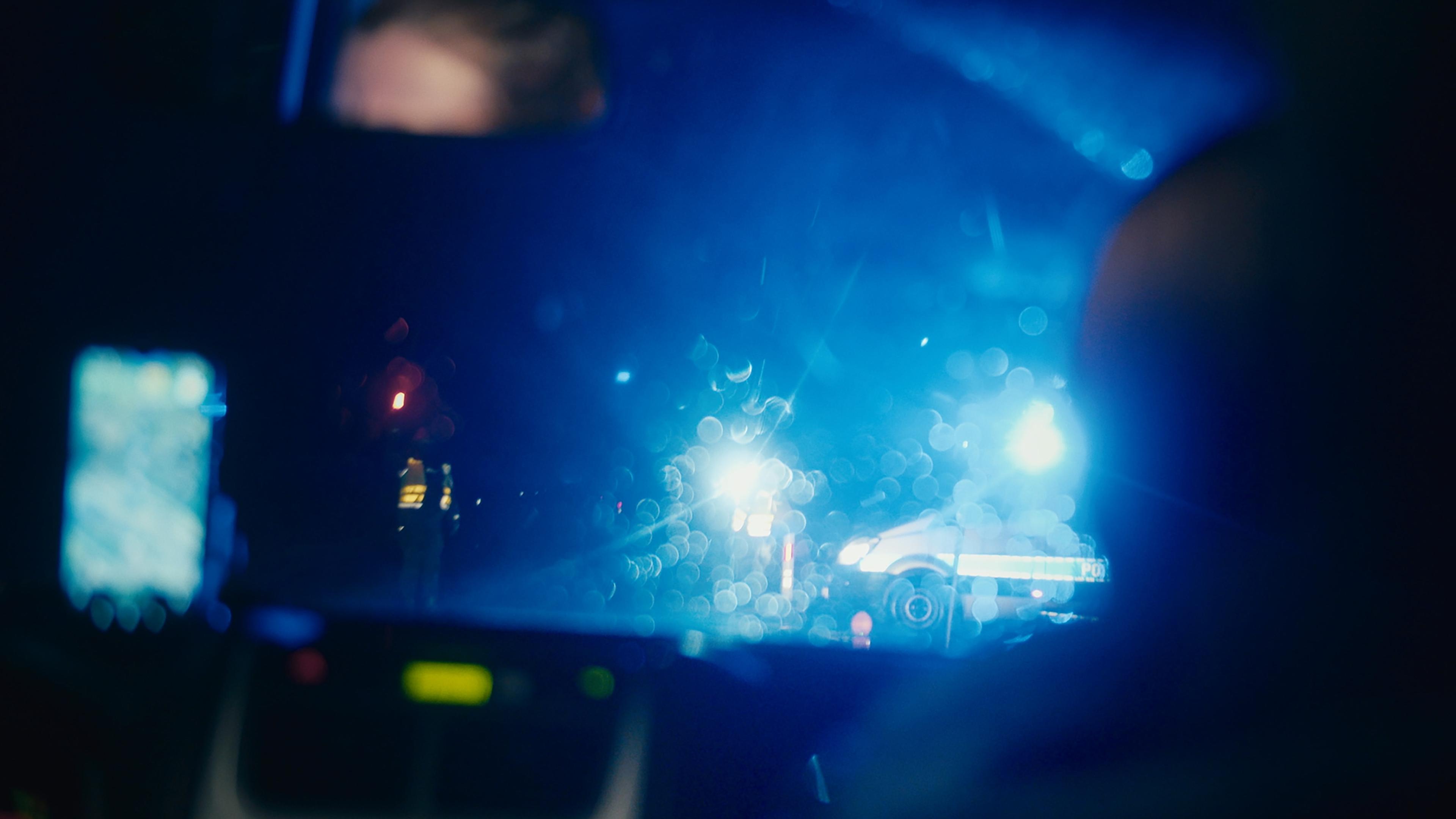 Blurry night photo through car window showing police lights and a figure, creating a dramatic scene with blue lighting.