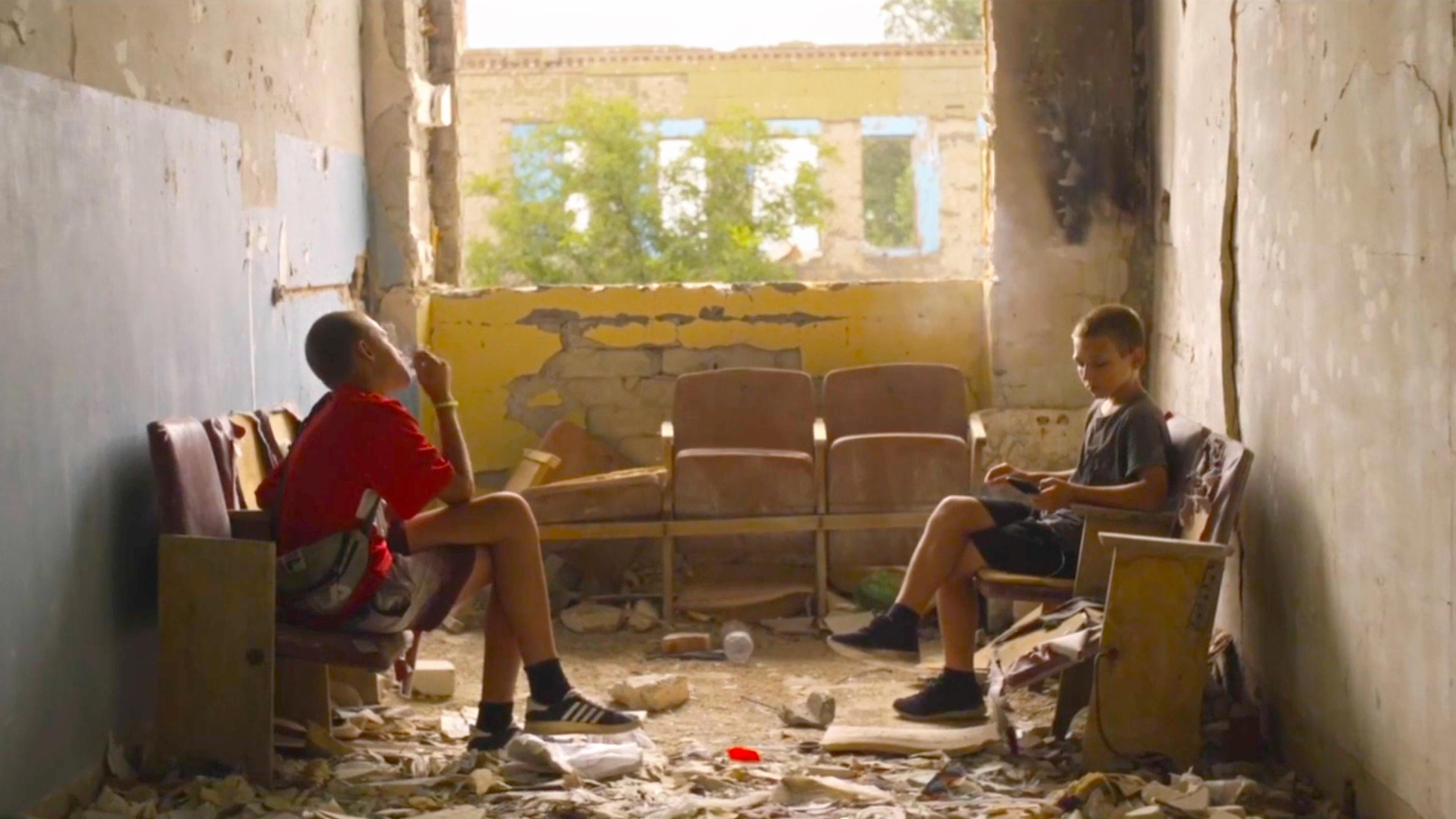 Photo of two boys sitting in a dilapidated room surrounded by debris with a view of an overgrown outside through a broken wall.
