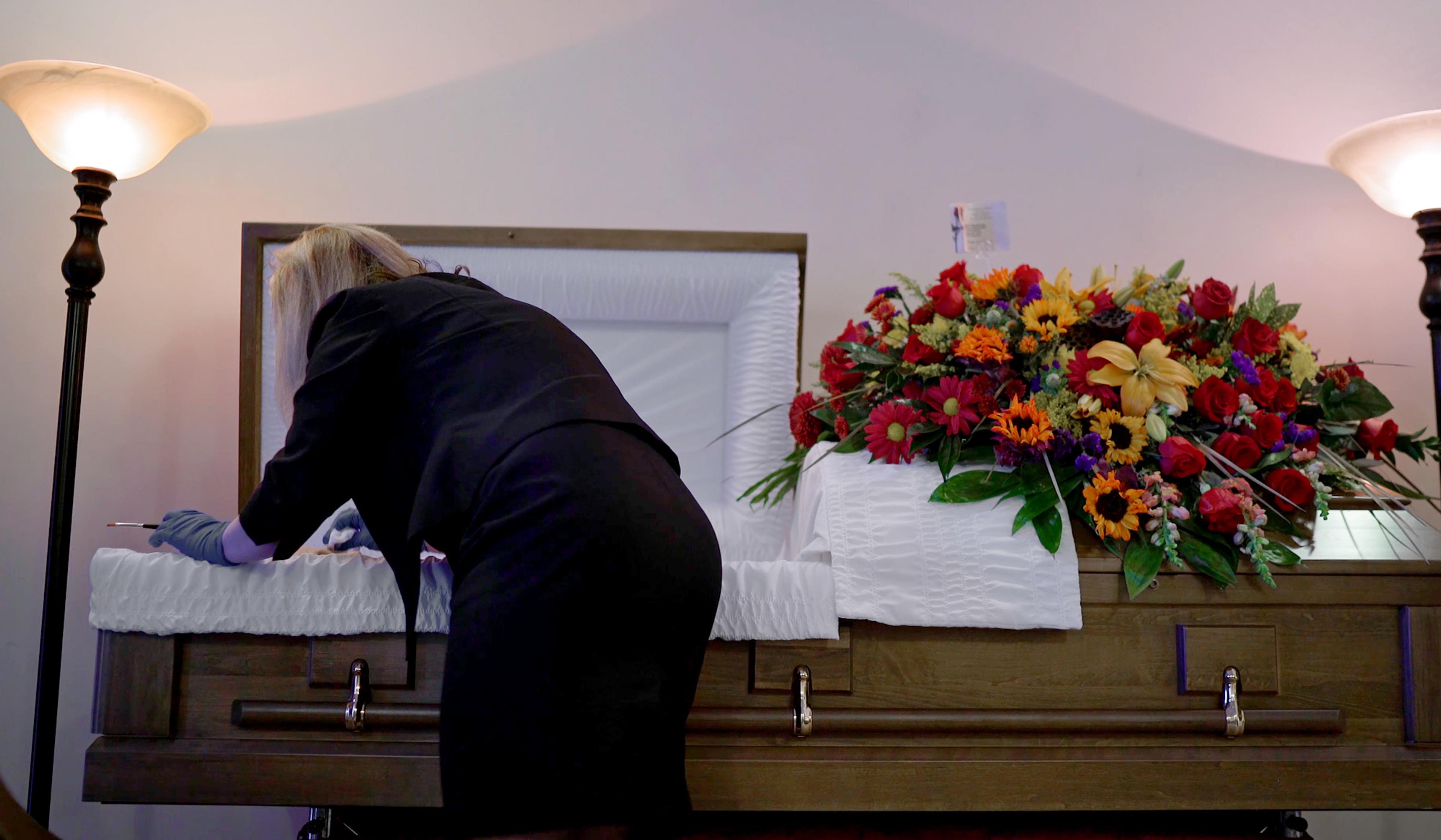 A person in black clothing and rubber gloves preparing a coffin adorned with a floral arrangement.
