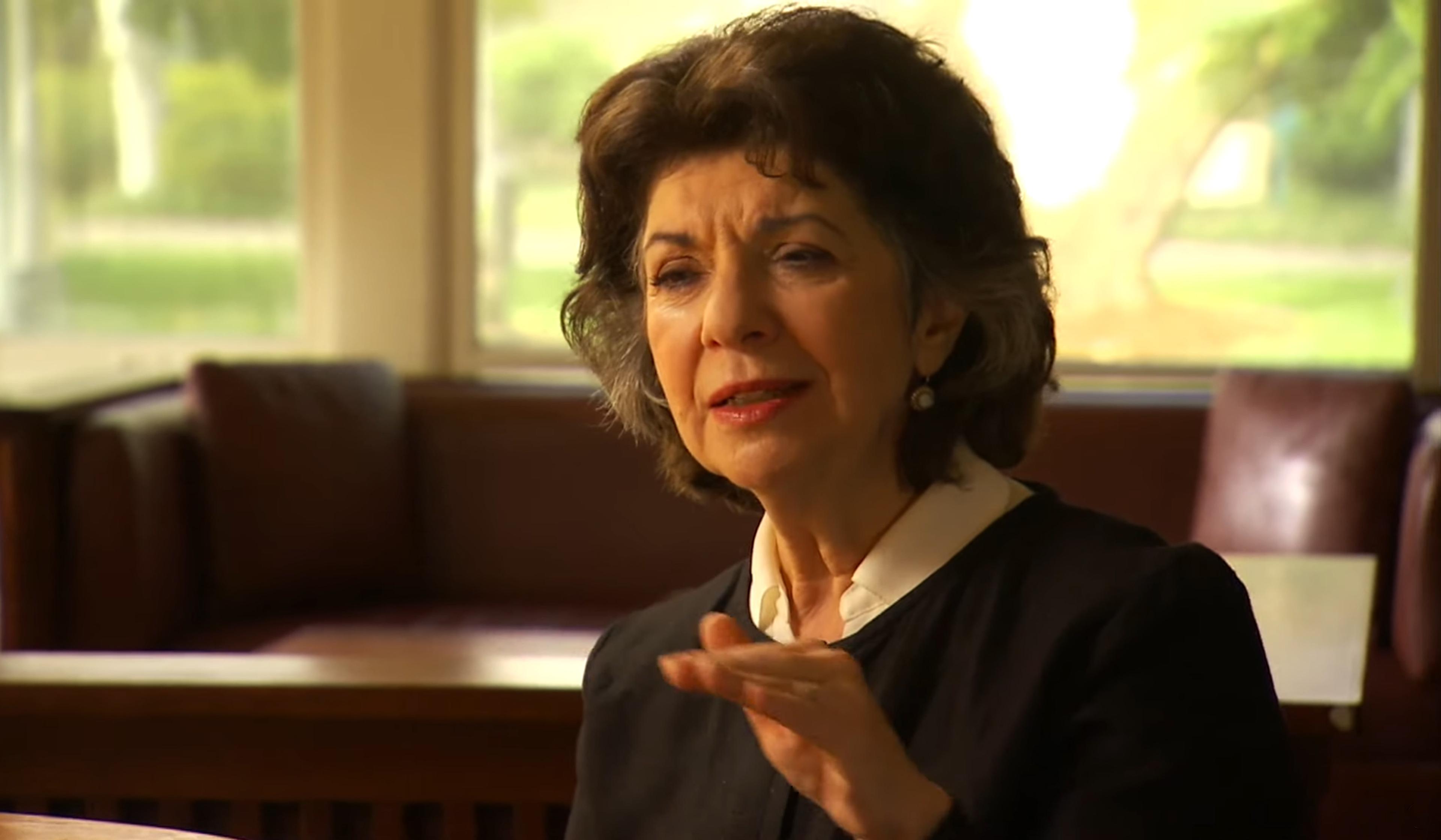 Elderly woman with short hair, wearing a black jacket and white shirt, speaking inside a room with large windows and blurred background.