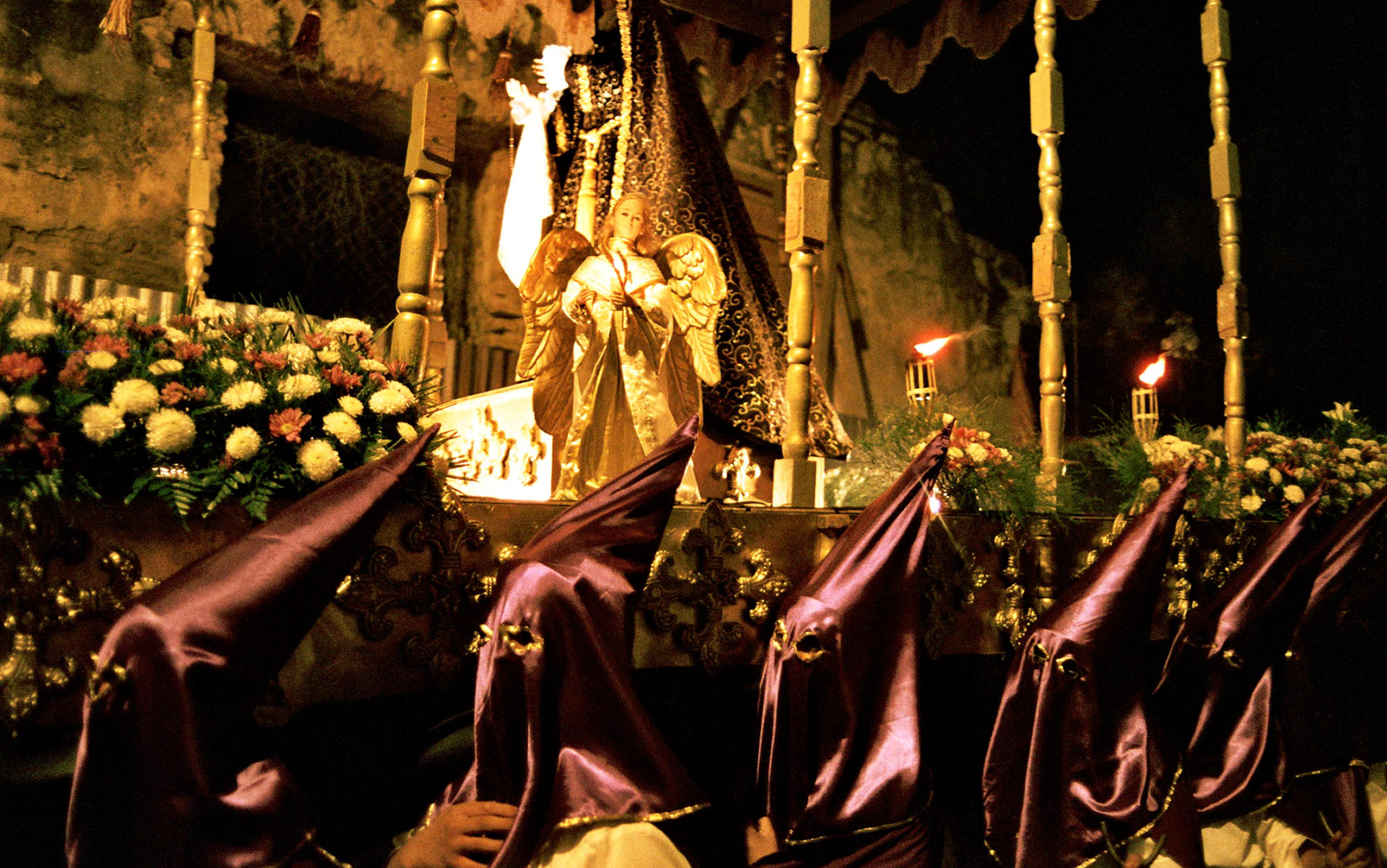 A religious procession with masked people in purple hoods carrying a statue adorned with angel figures and flowers at night.