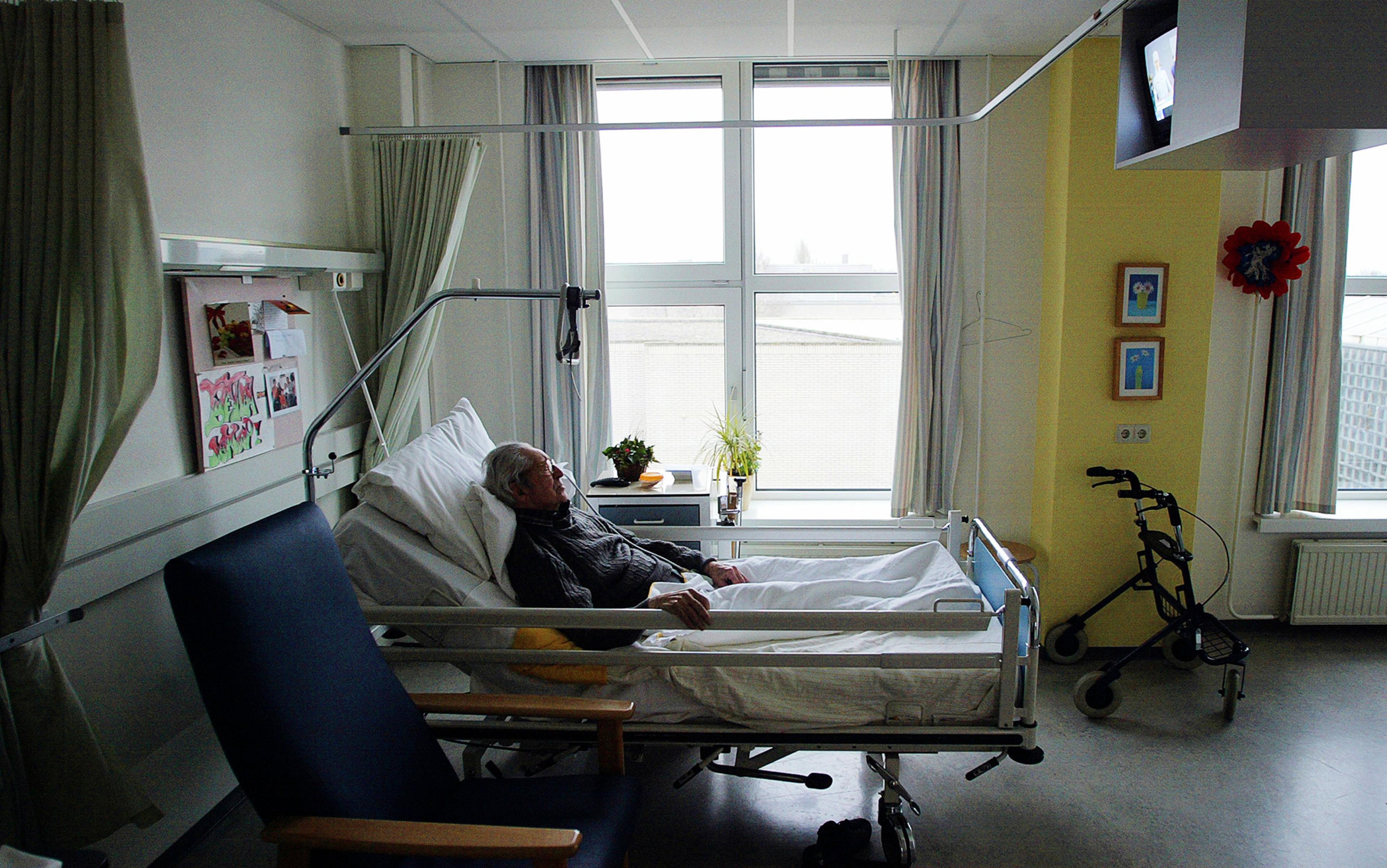 An elderly person lying in a hospital bed in a bright room with a walker and a window nearby.