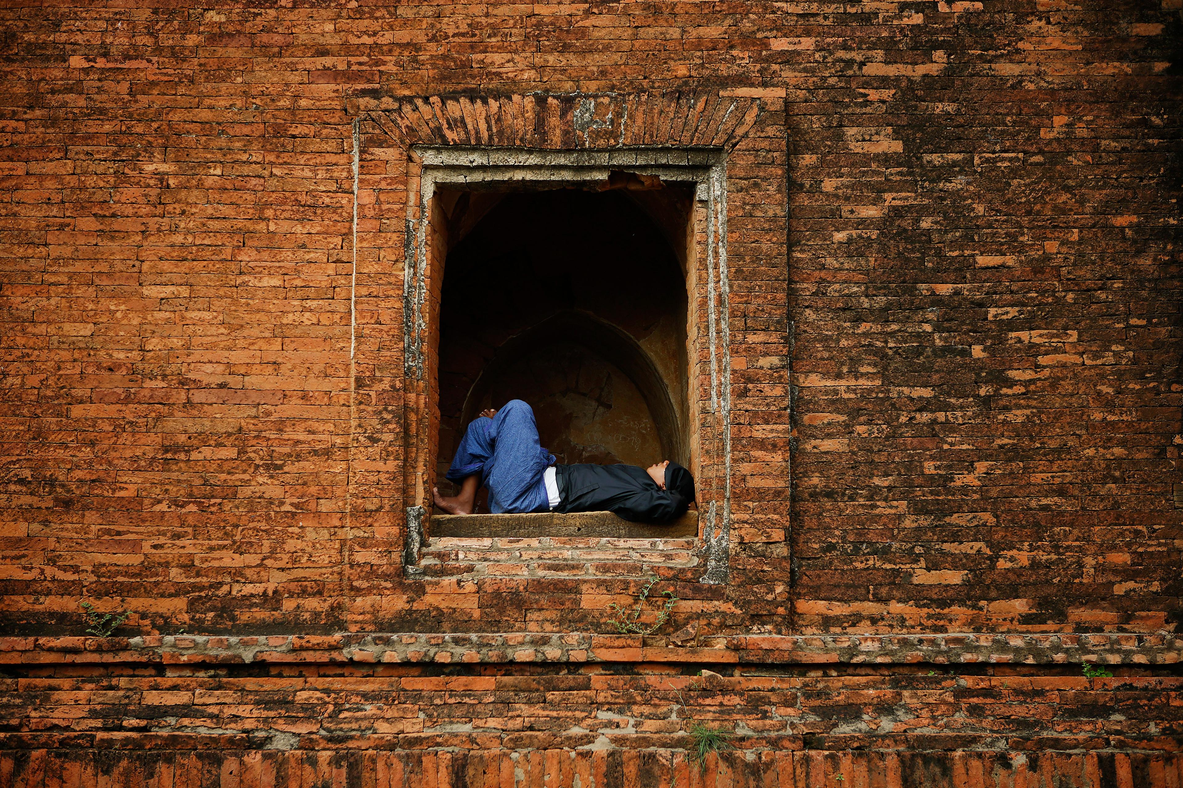 A person in blue jeans and black jacket resting in an alcove in an old red brick wall.