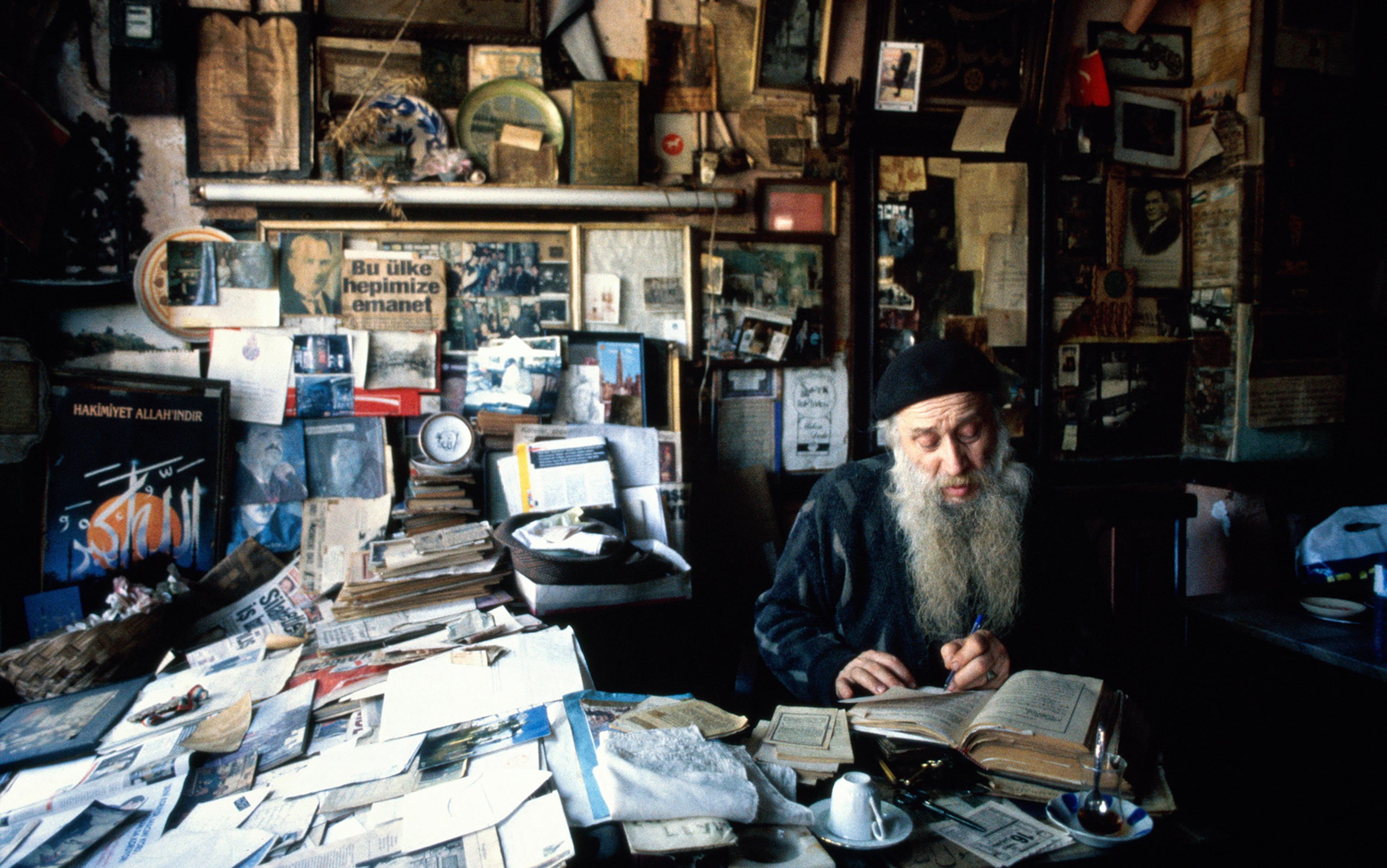 An elderly man with a long beard writing at a cluttered desk in a room filled with papers, books and various memorabilia.