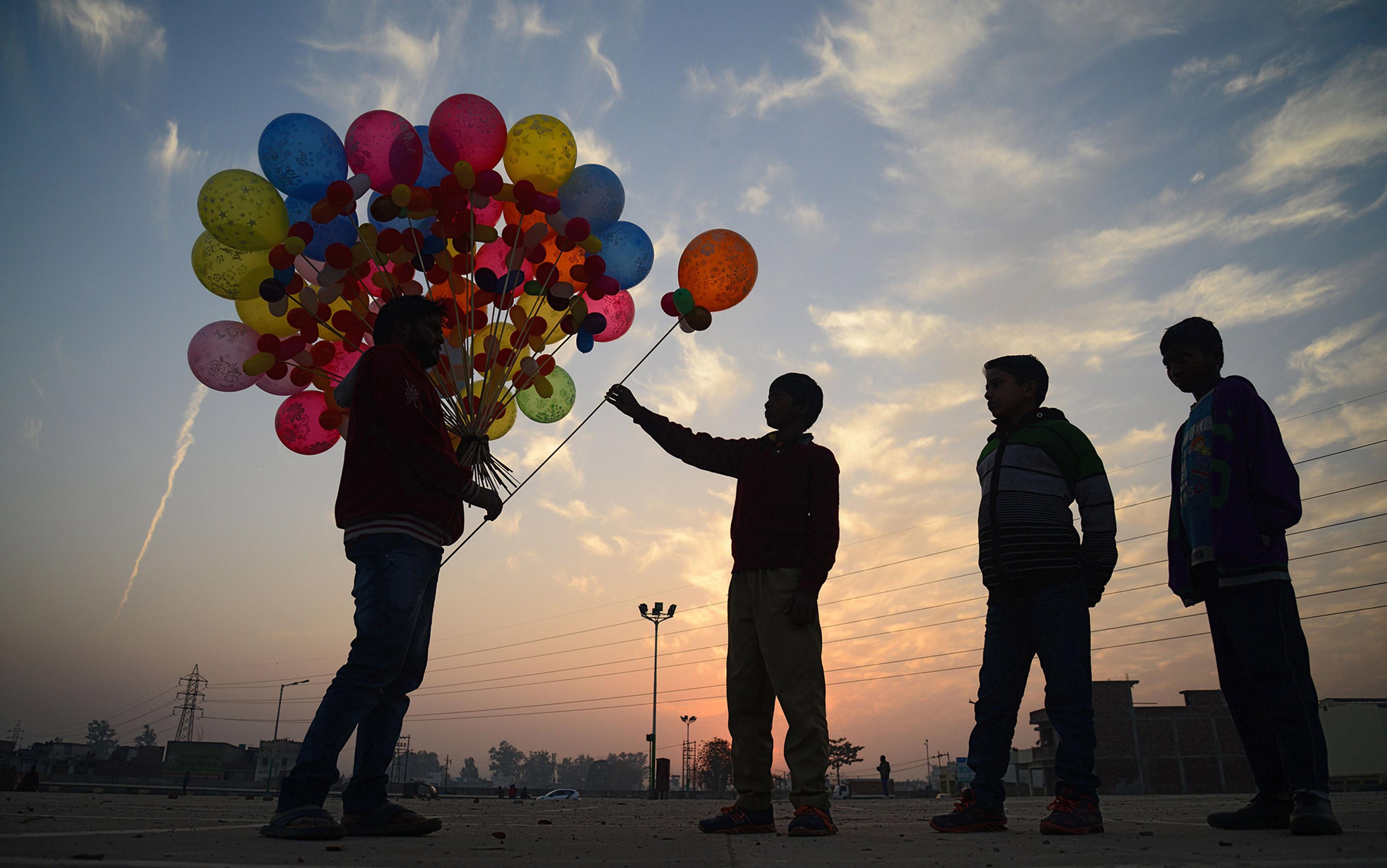 Three silhouetted children at sunset, with one child buying colourful balloons from a seller in an open area with a cloudy sky.