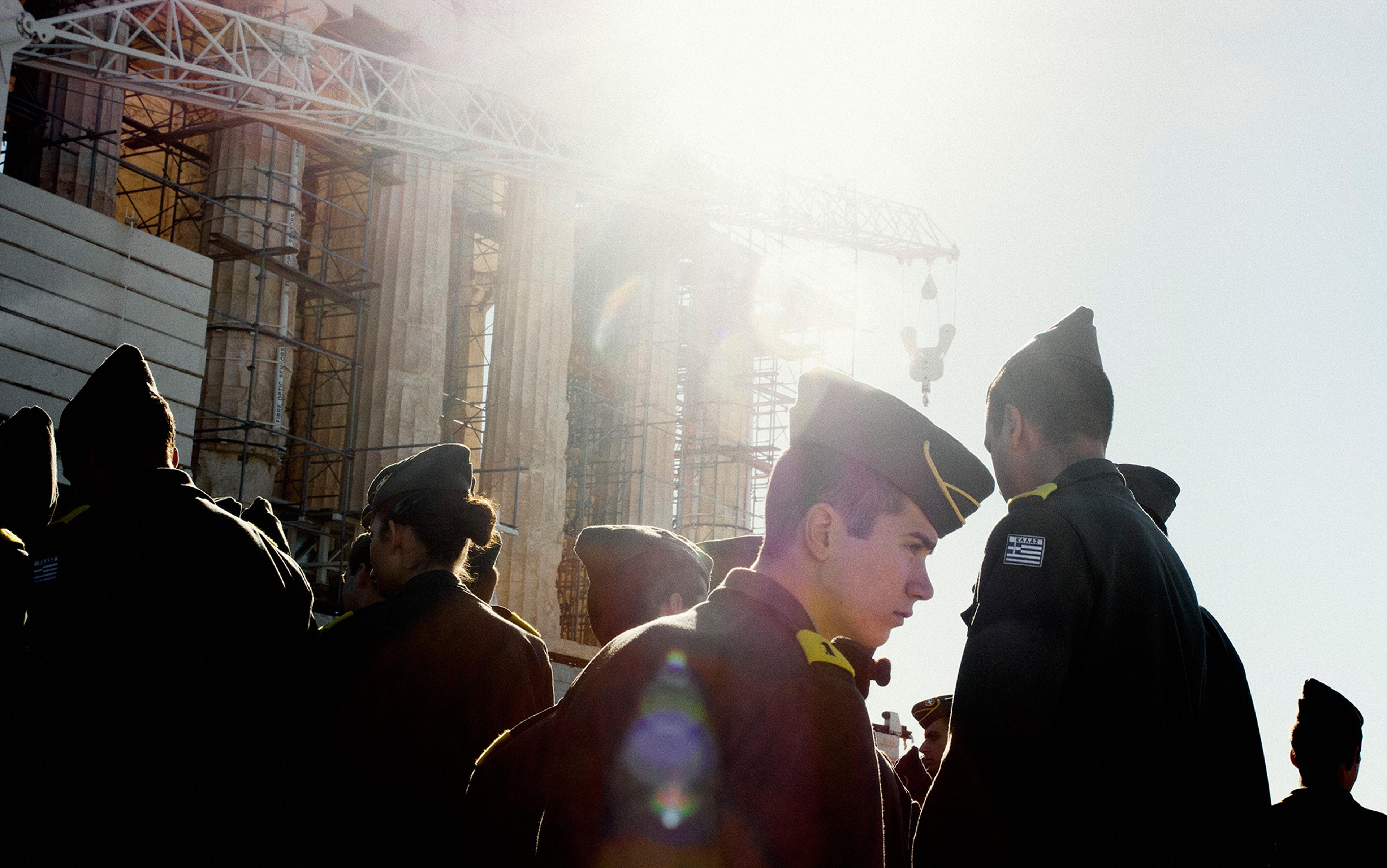 A group of uniformed individuals wearing the Greek flag insignia, backlit by the sun, in front of a construction site with scaffolding and ruins.