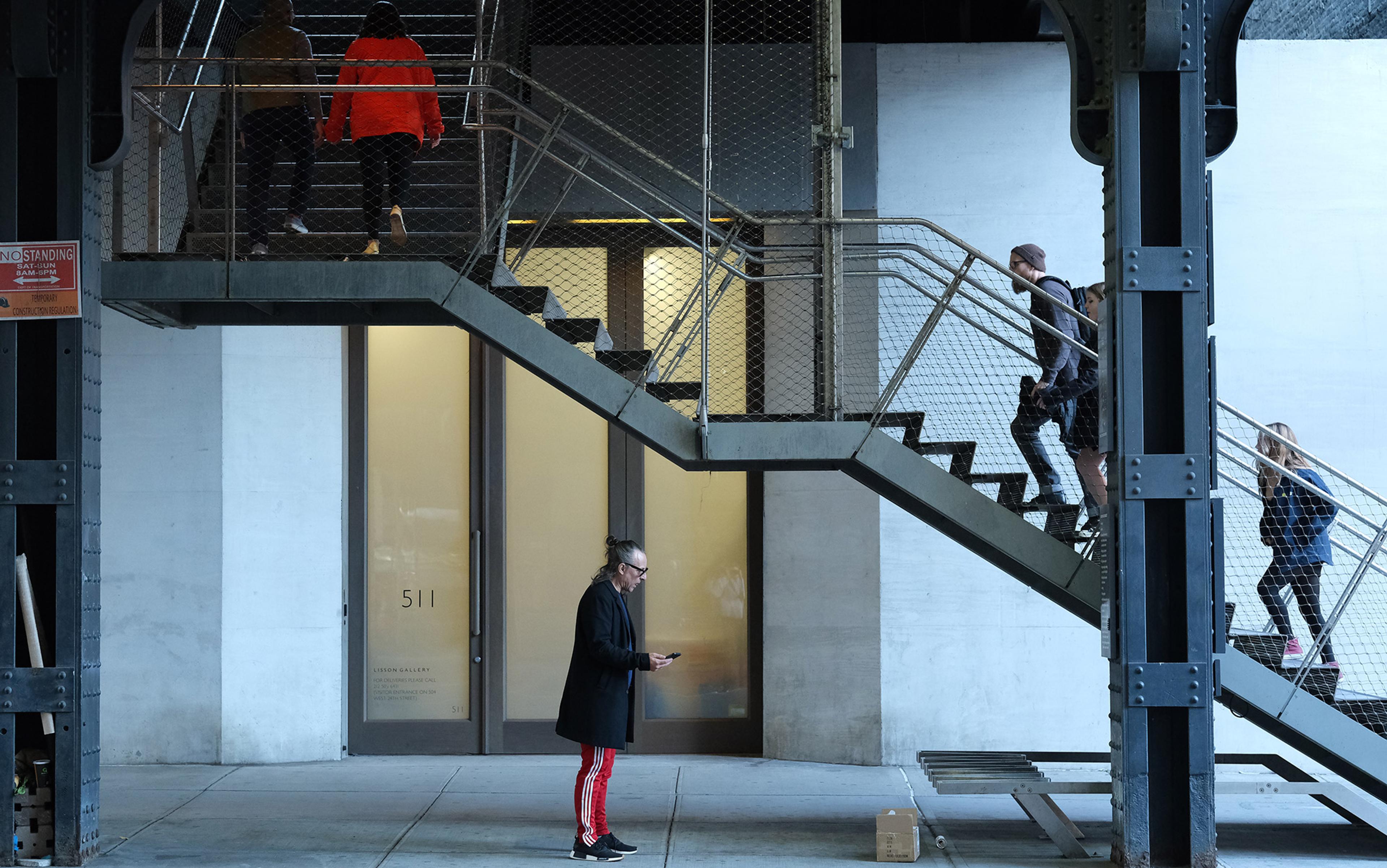 People walking on stairs inside a modern industrial building, with someone standing below checking their phone; entrance to Lisson Gallery visible in the background.