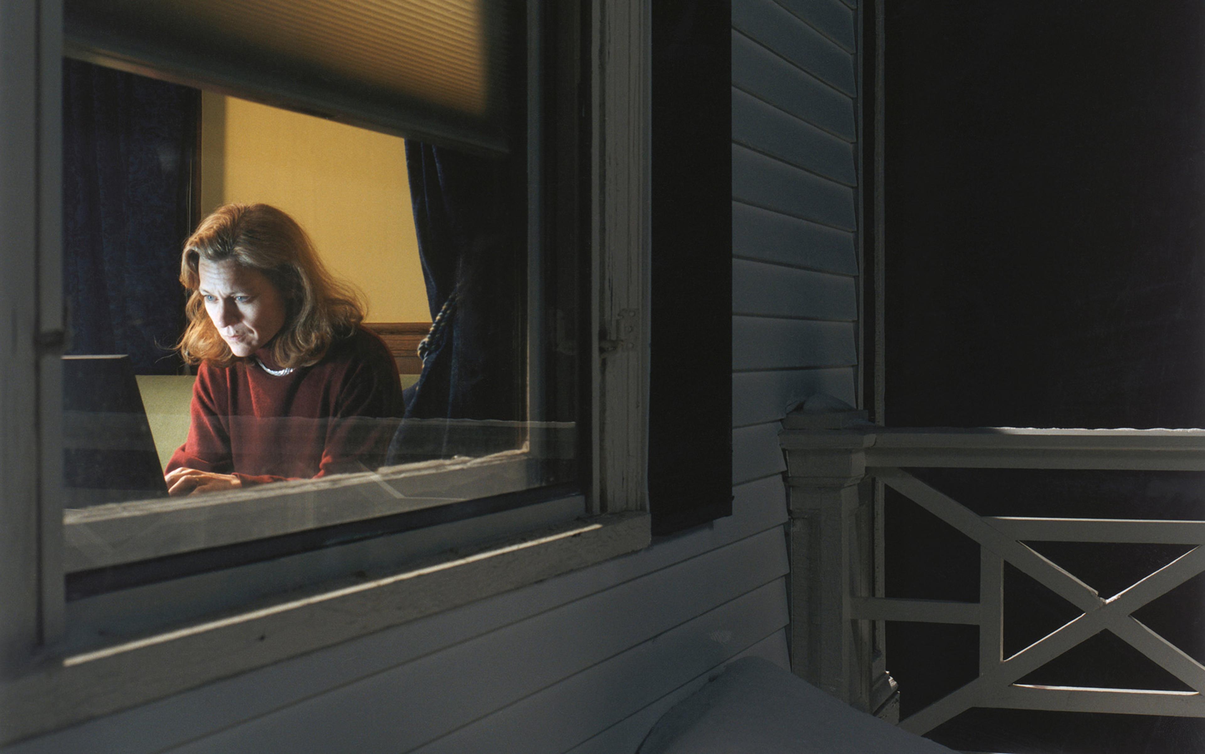 A woman working on a laptop indoors at night viewed from outside through a window, with dark curtains and a dimly lit room.