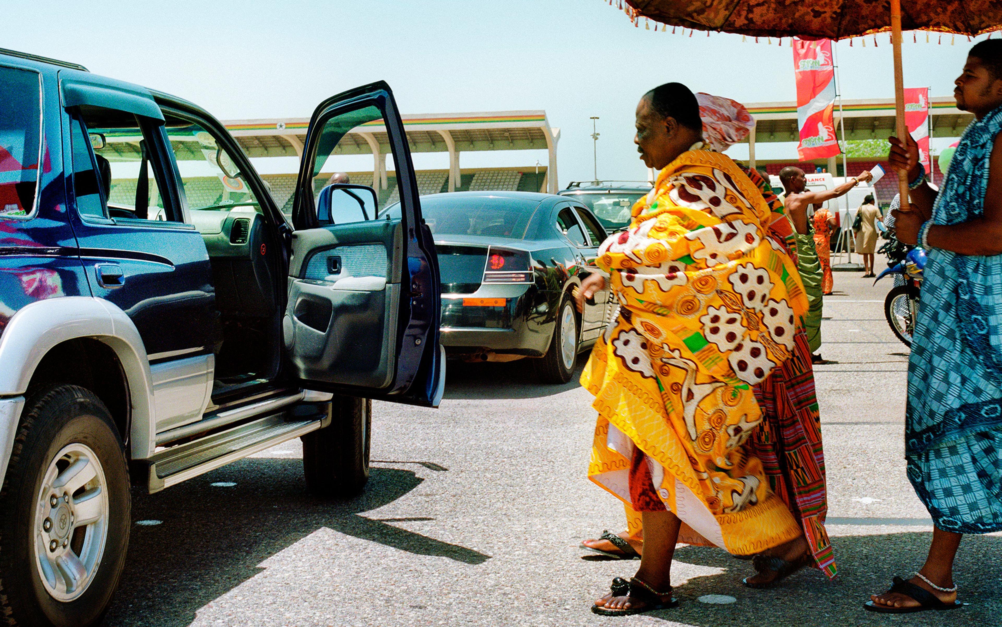 A man in vibrant traditional African attire approaching an open car door, with a stadium and other people in the background.