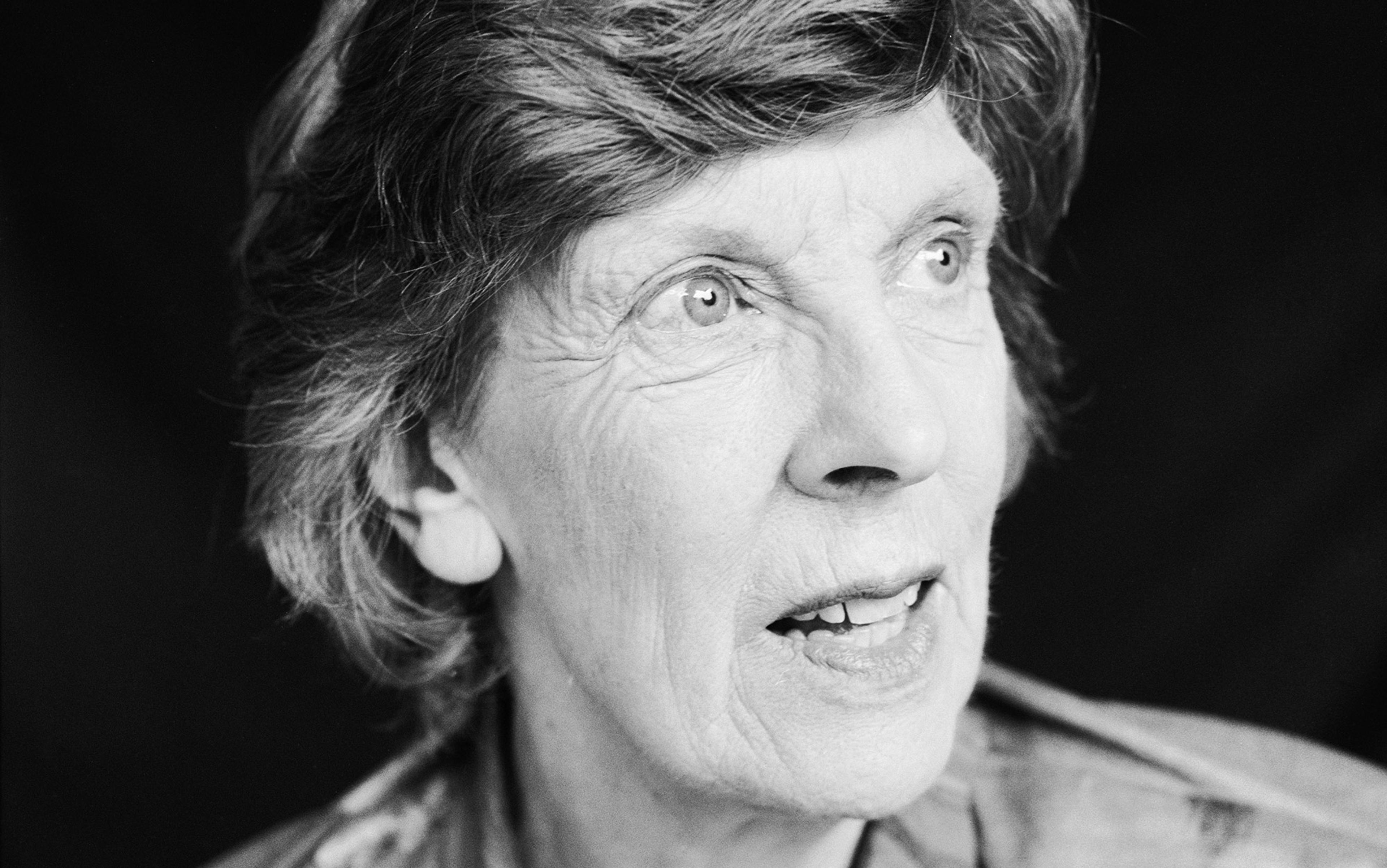 Black and white photo of an older woman with short hair looking to the side with a thoughtful, open expression and a dark background.