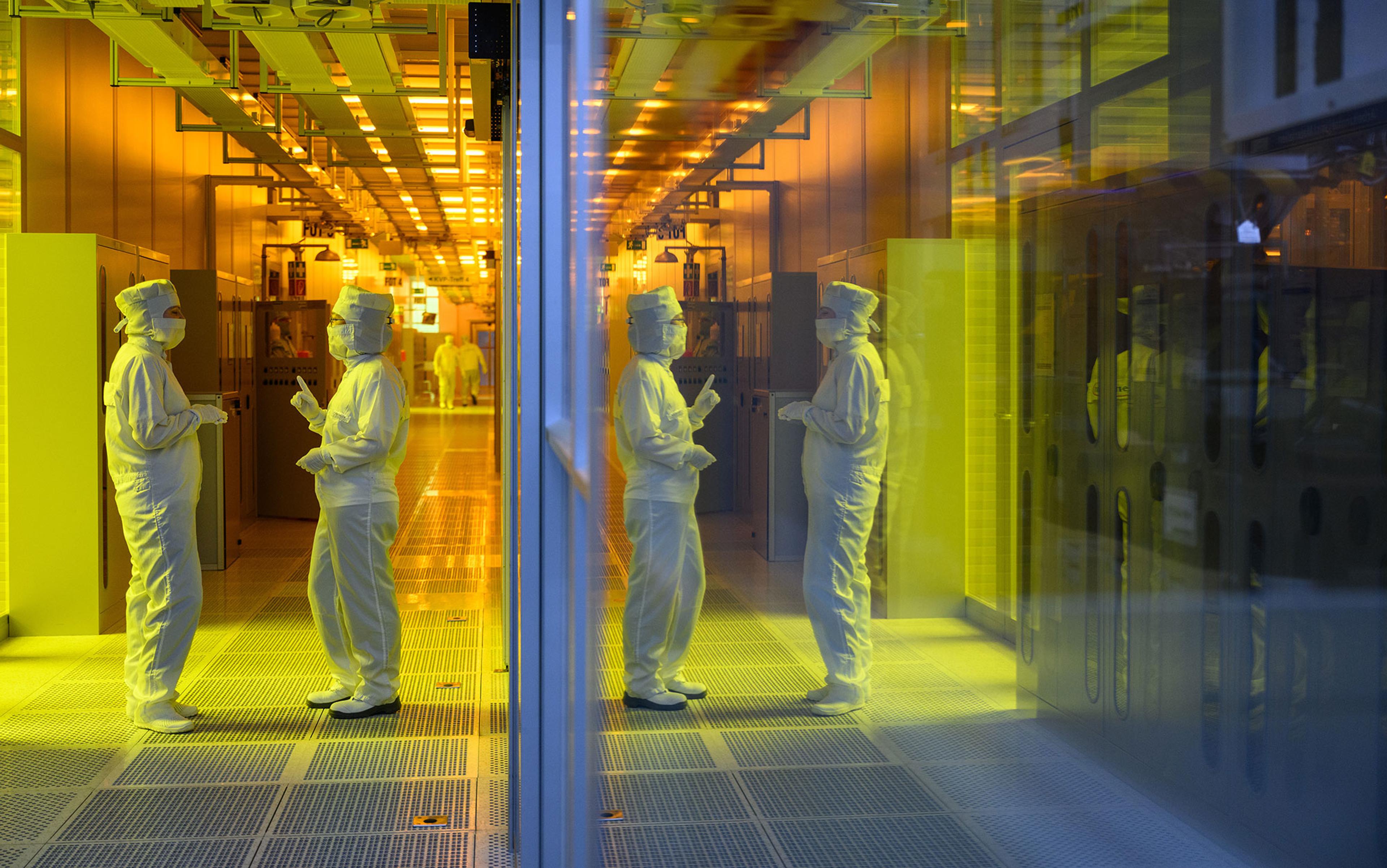 Scientists in cleanroom suits conversing in a brightly lit laboratory with a yellow hue, featuring reflective surfaces and specialised equipment.