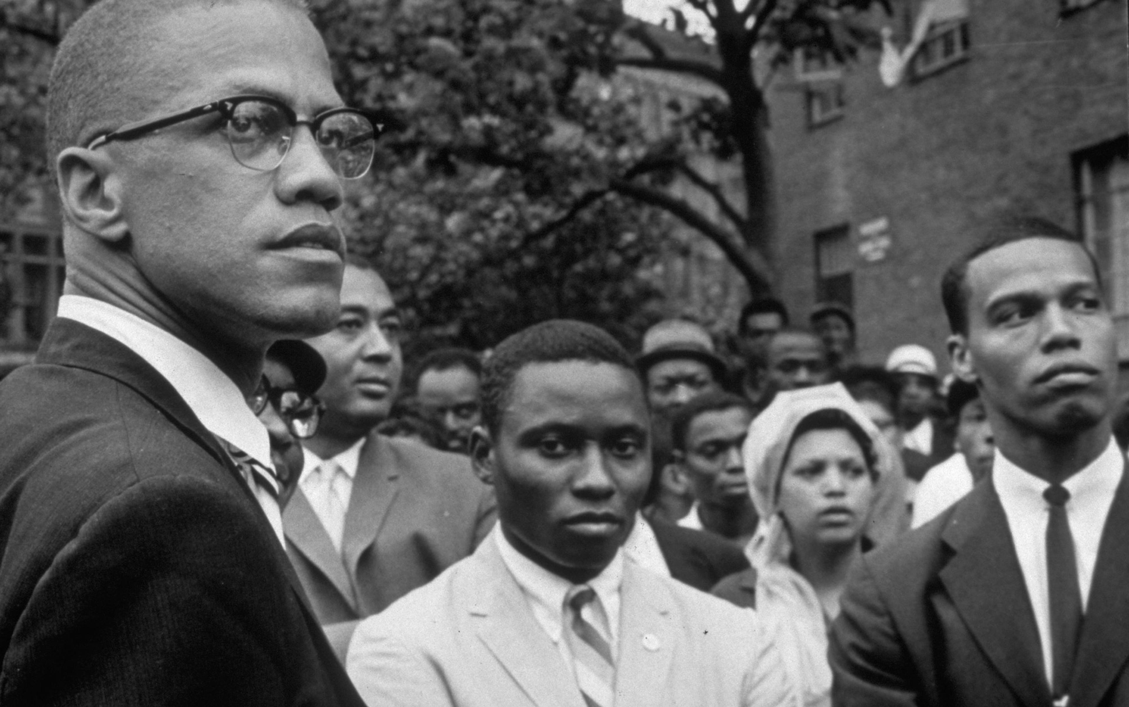 Vintage photo of a Black man with glasses who is Malcolm X and a crowd in formal attire, standing outdoors near buildings and trees.