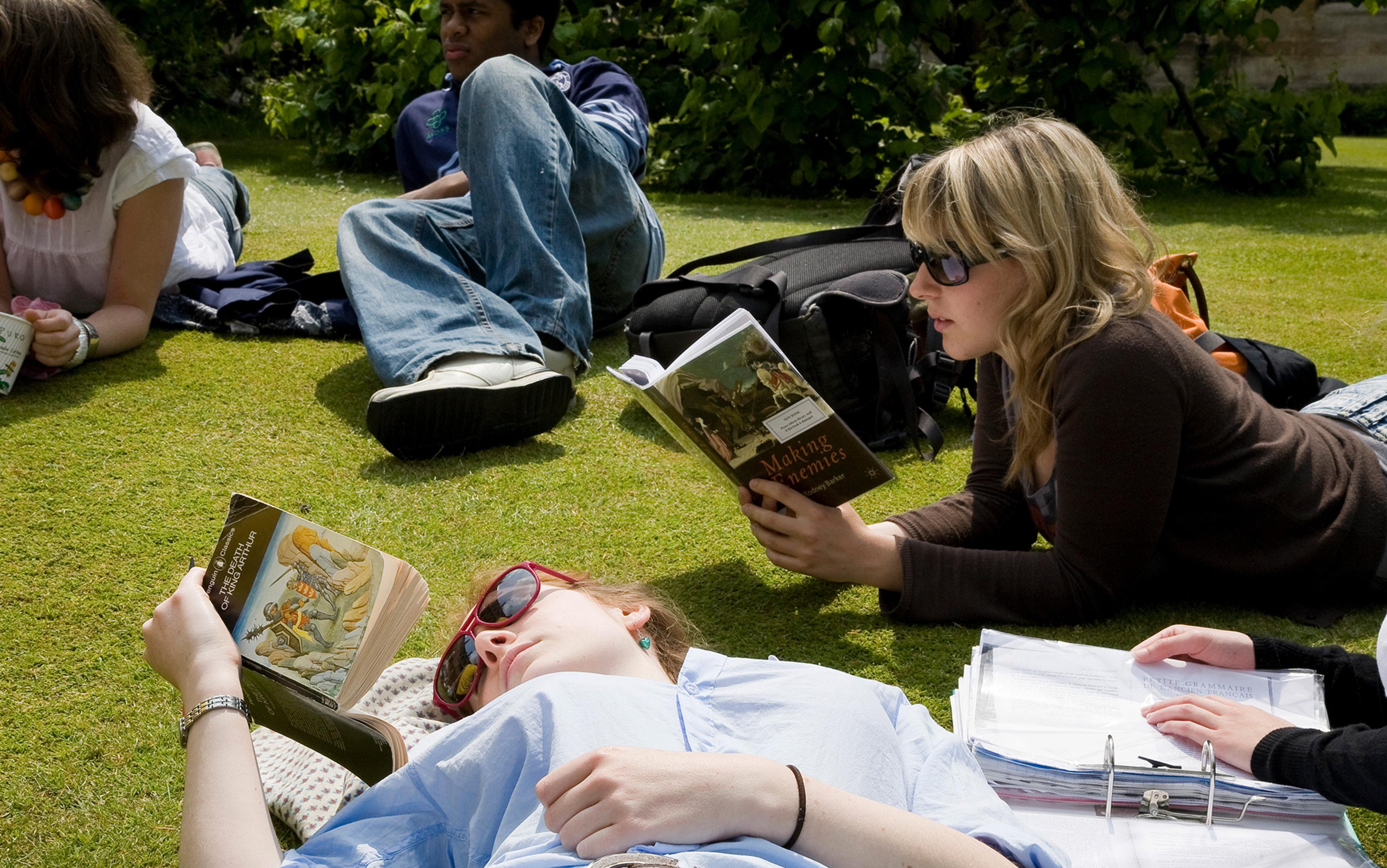 Young people lying on grass, reading books and studying, with backpacks and notes around them, on a sunny day.