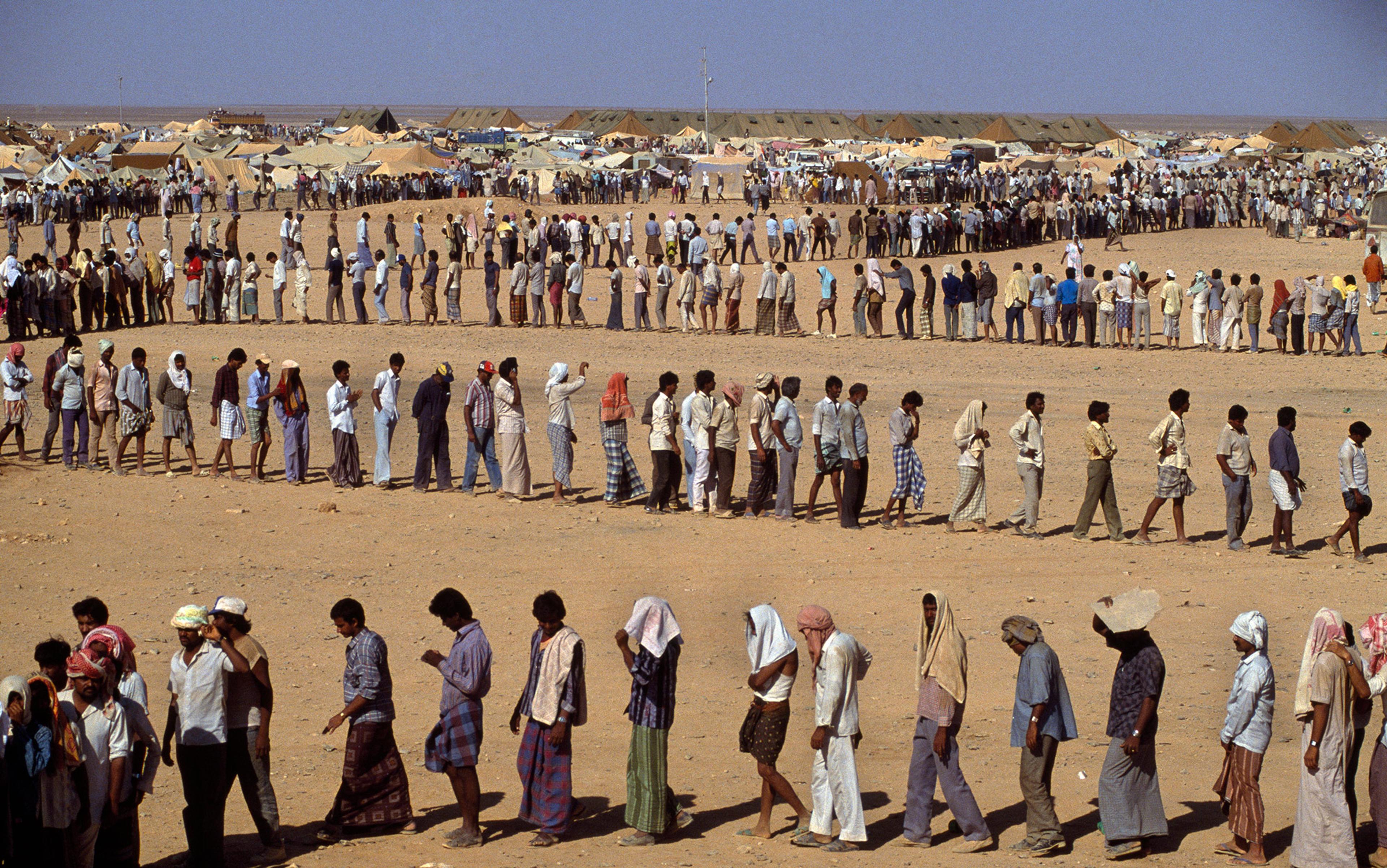 People standing in long, winding queues in a desert camp with numerous tents in the background under a clear blue sky.