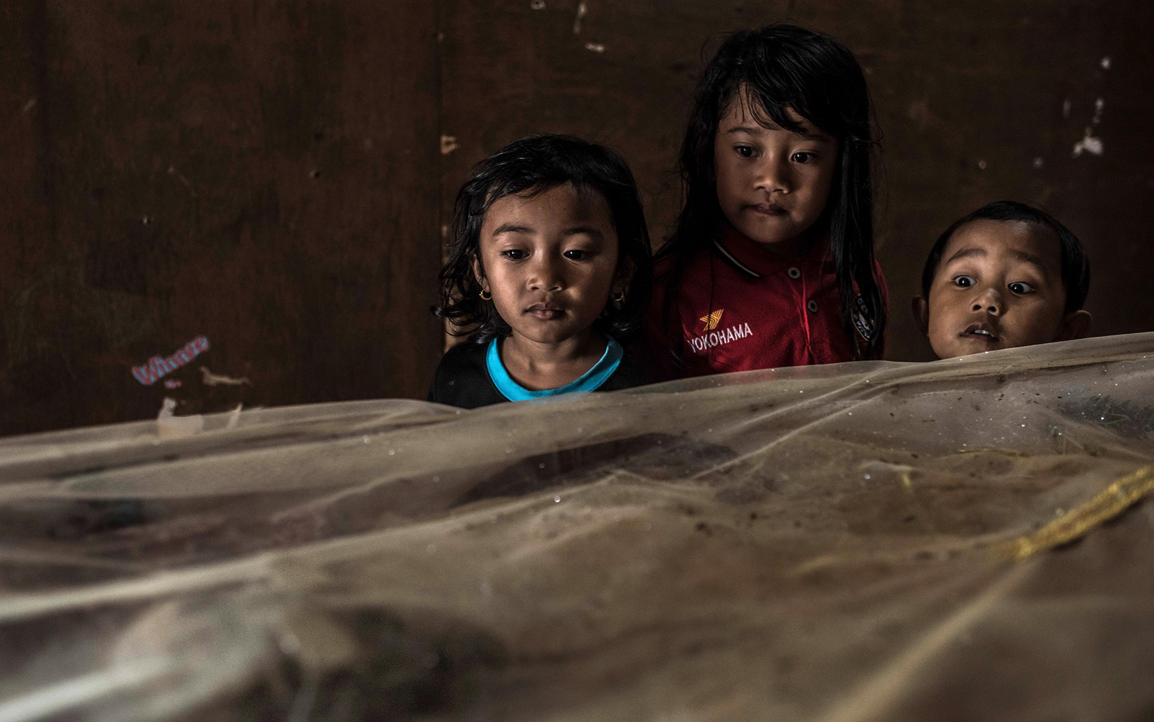 Three children looking curiously at an object covered with a translucent cloth in a dimly lit room.