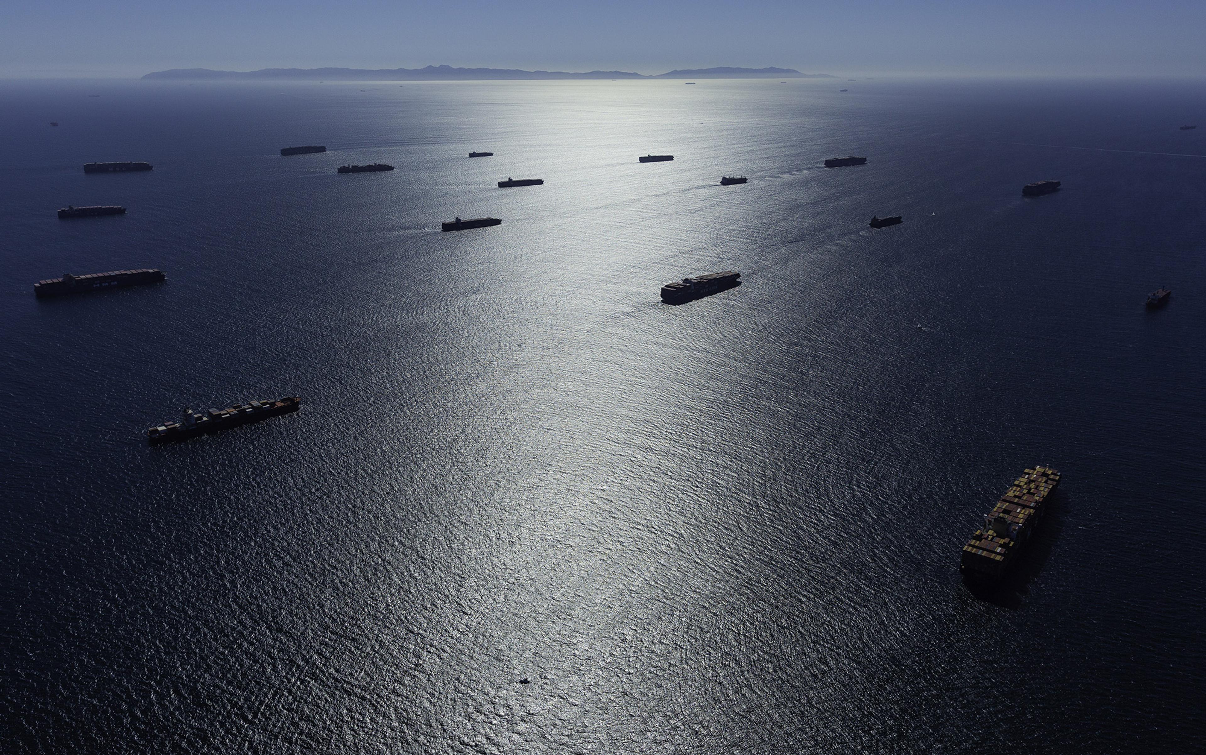 Aerial view of numerous container ships on a shimmering sea with distant mountains under a clear sky.