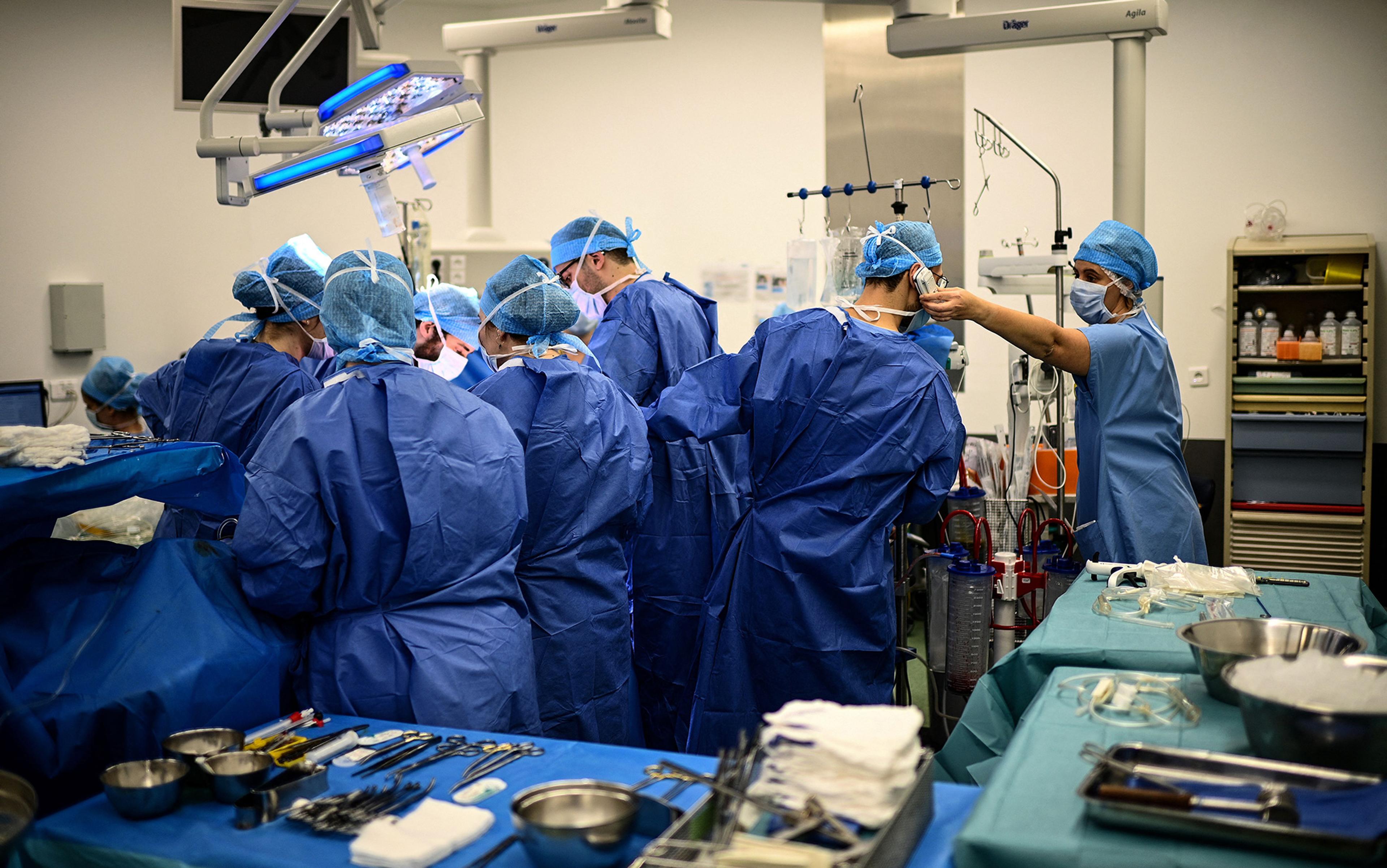 A team of surgeons in blue scrubs and surgical caps operate under bright surgical lights in an operating theatre. Medical instruments and equipment are arranged on tables around them, while one team member adjusts another’s mask. Shelves in the background hold medical supplies.