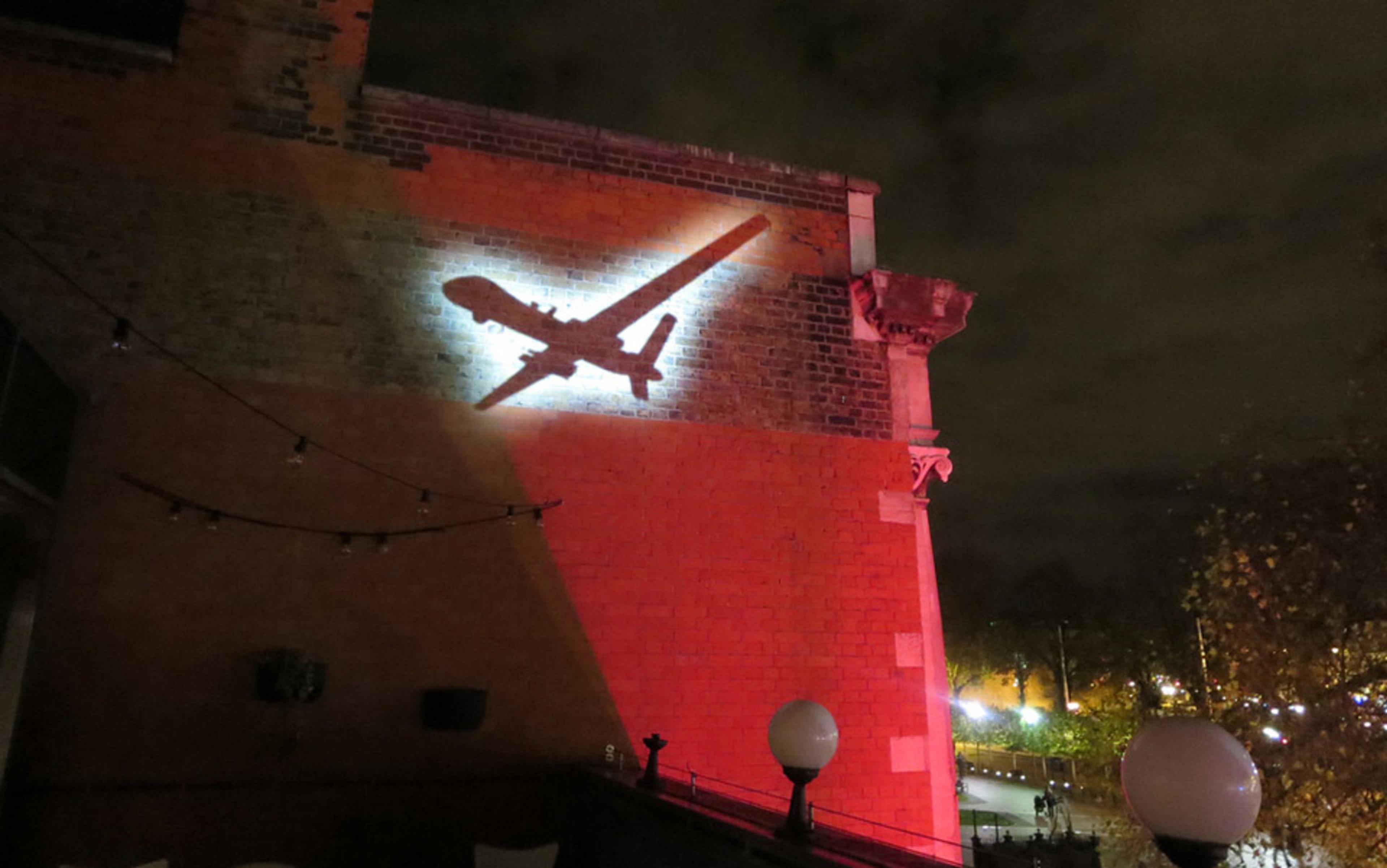 A brick wall with a projected shadow of an aeroplane, surrounded by red lighting, with trees and lights in the background.