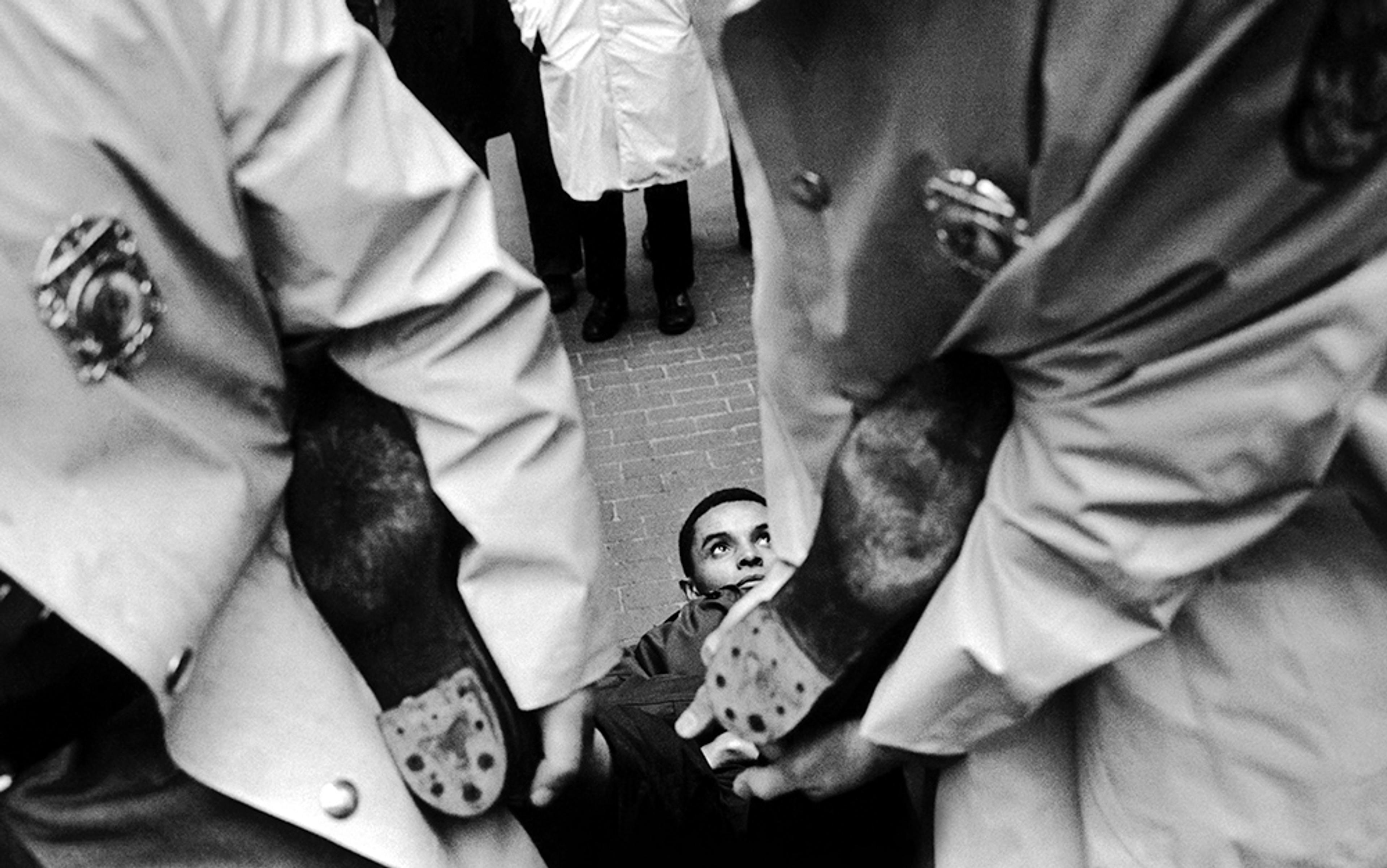 Black and white photo of a person being carried by two uniformed officers, viewed from below as onlookers stand in the background.