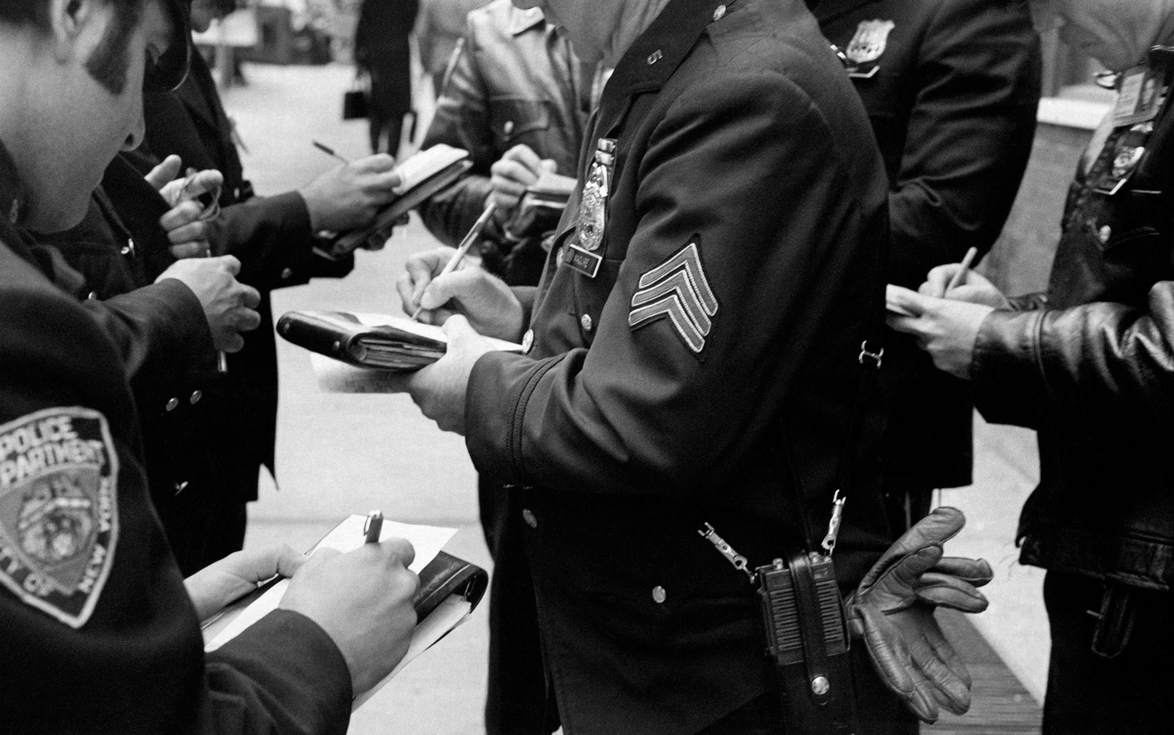 Black and white photo of police officers writing in notepads, focusing on their hands and uniforms with badges visible, in an outdoor setting.