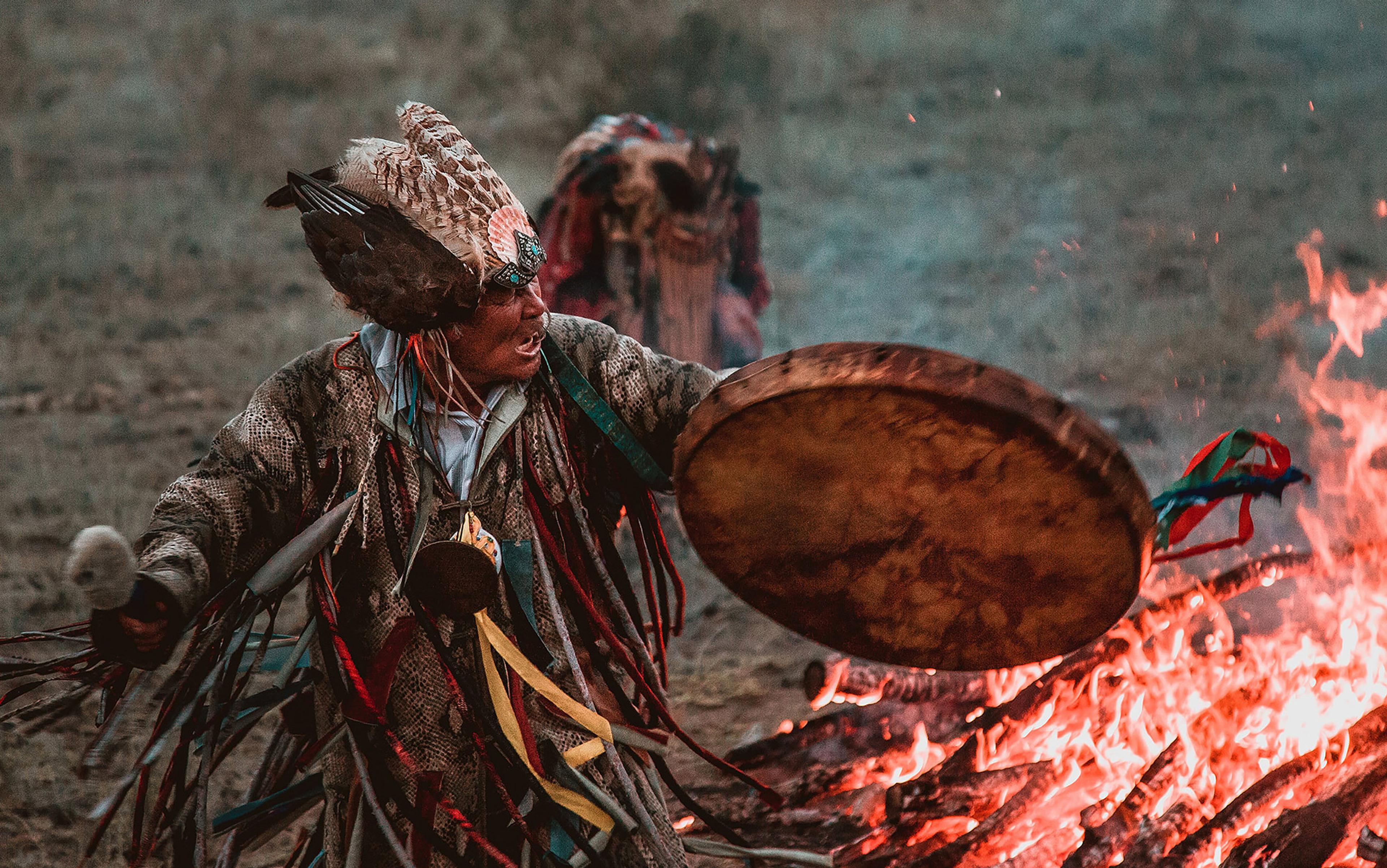 A person in tribal attire with feathers holding a drum by a fire, performing a ritual in an outdoor setting.