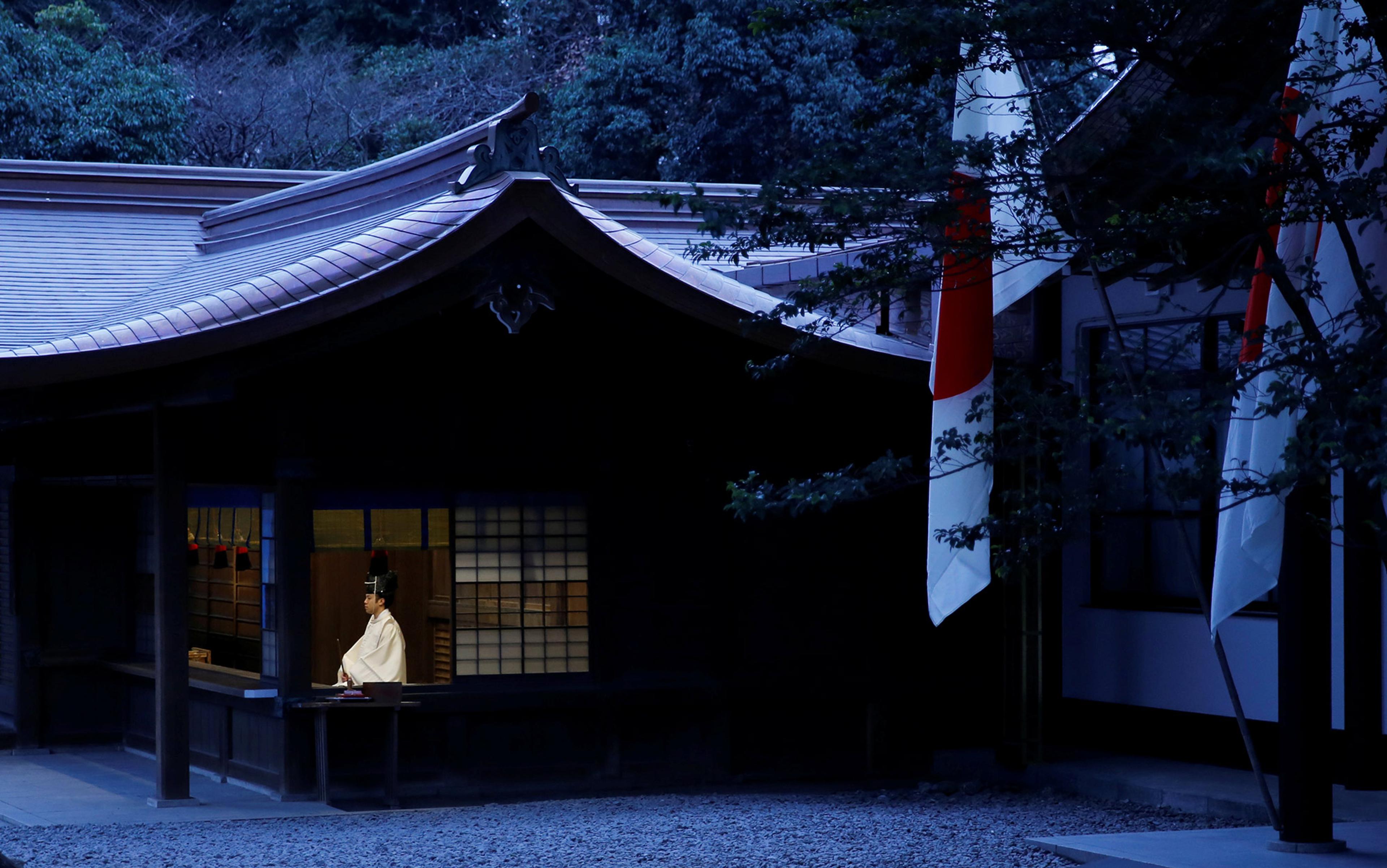A Japanese monk in traditional clothing on the porch of a building at night with large flags displayed outside next to trees.
