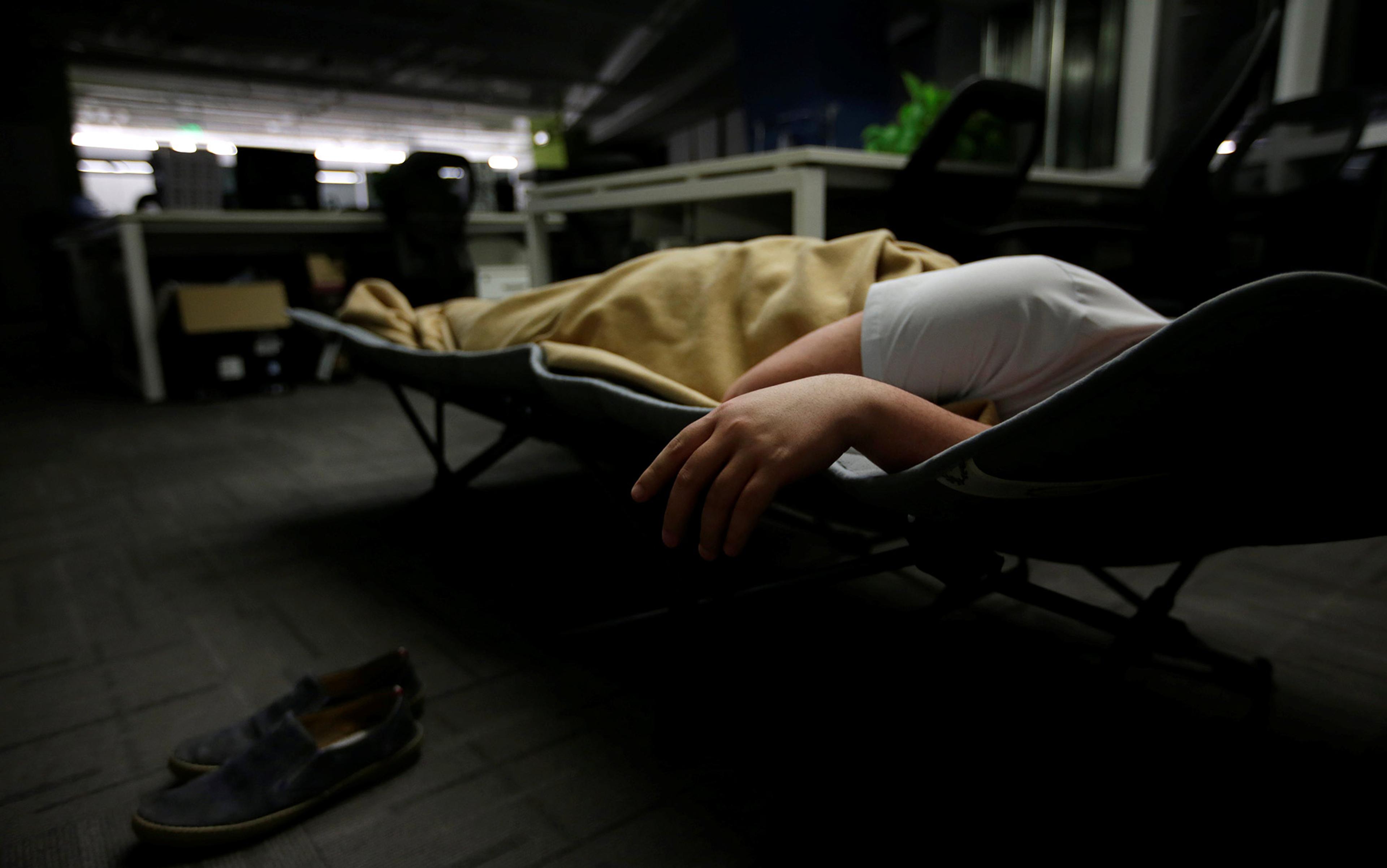A person sleeping on a folding cot in an office, wearing a white shirt. Shoes are placed on the carpeted floor nearby.