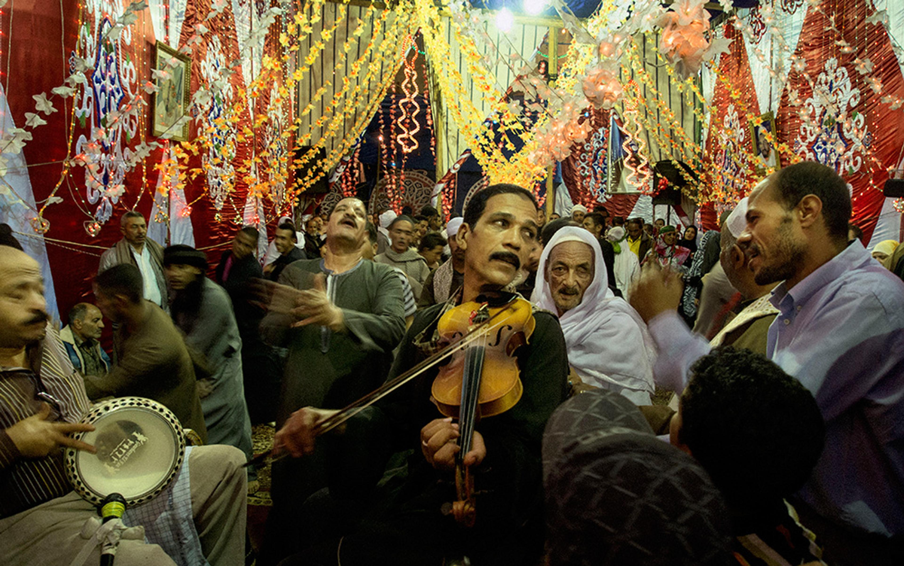 A lively celebration with musicians, people dancing, and colourful decorations hanging from the ceiling in a festive room.