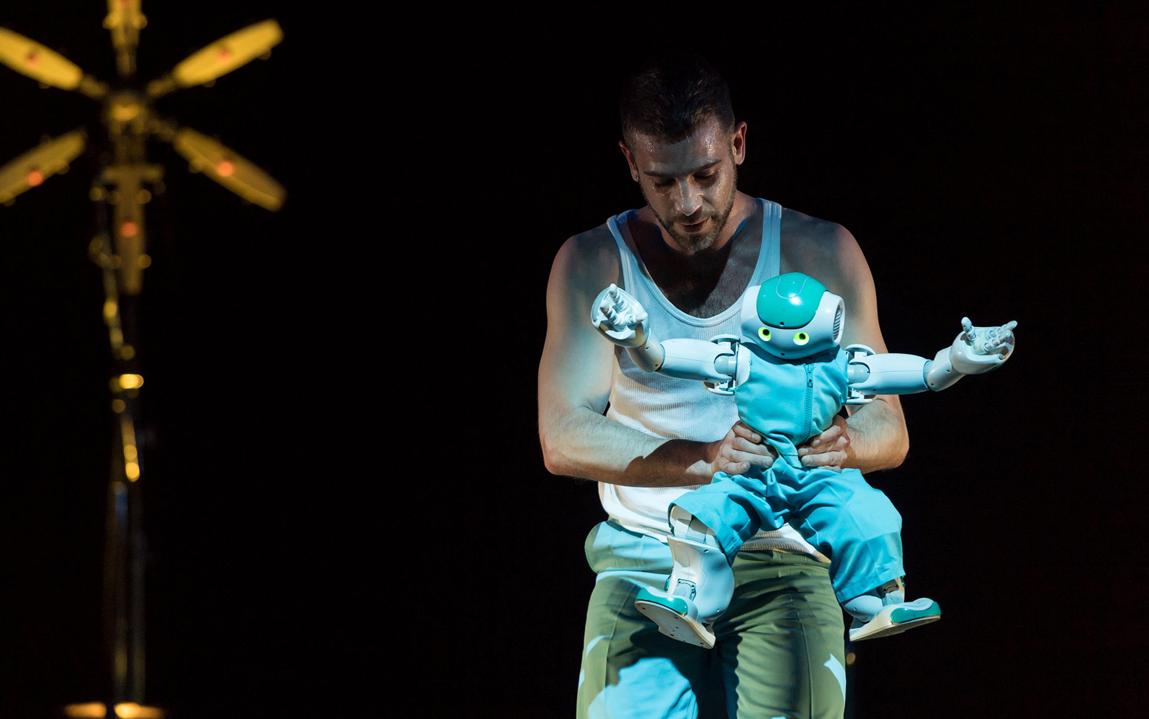 A man in a white vest holding a small, humanoid robot in blue clothing against a dark background with a yellow light.