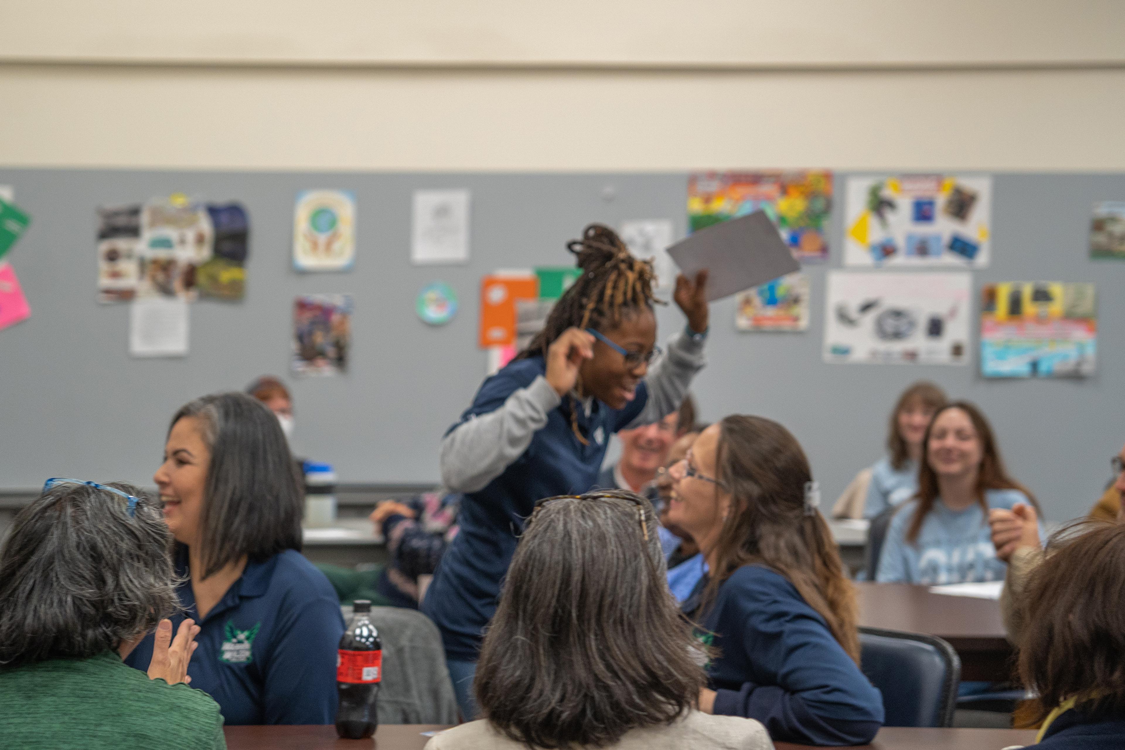 A lively classroom with smiling people, one person standing while holding a paper, posters in the background.