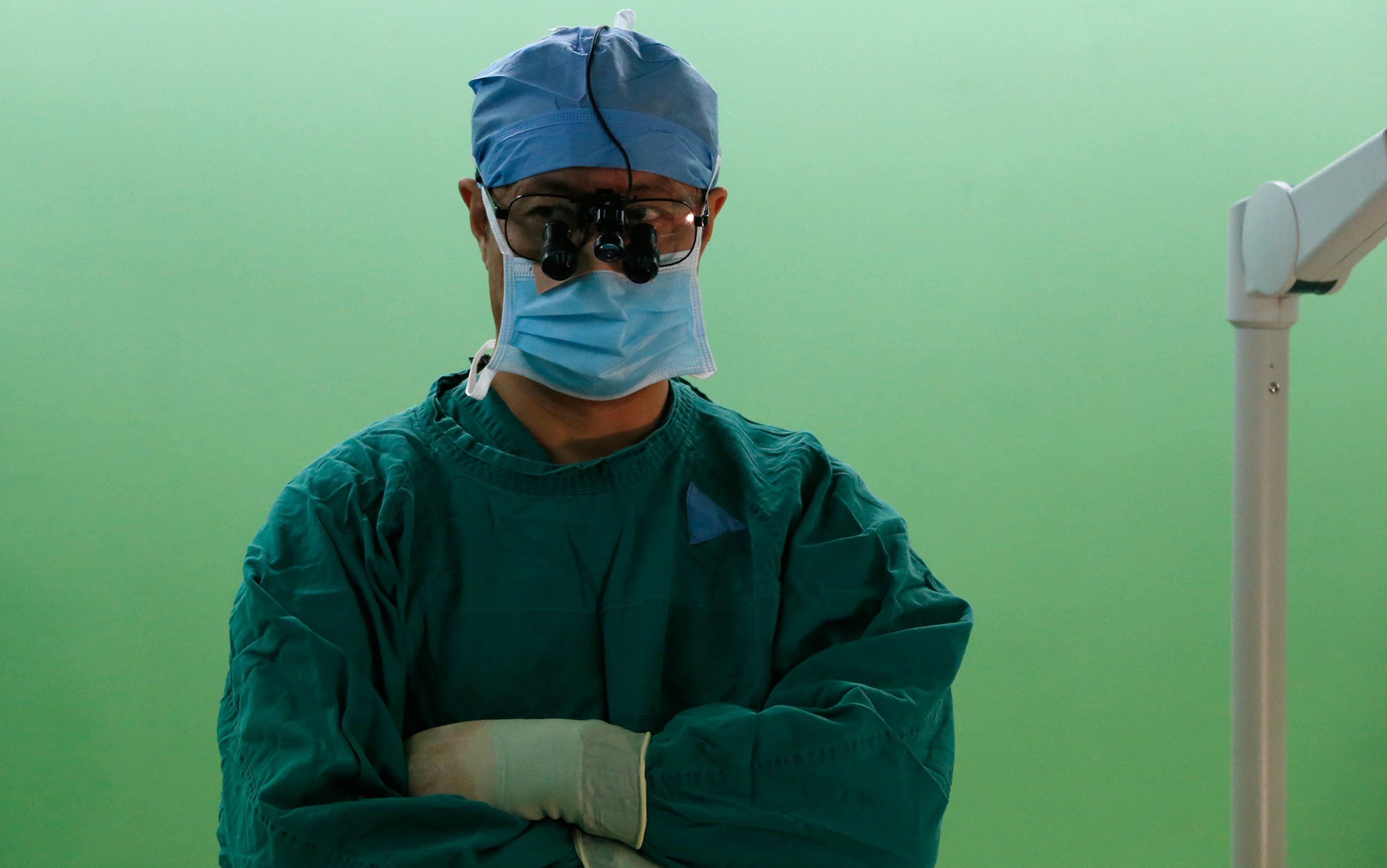 A surgeon in scrubs, face mask, and magnifying glasses standing with arms crossed against a green background.