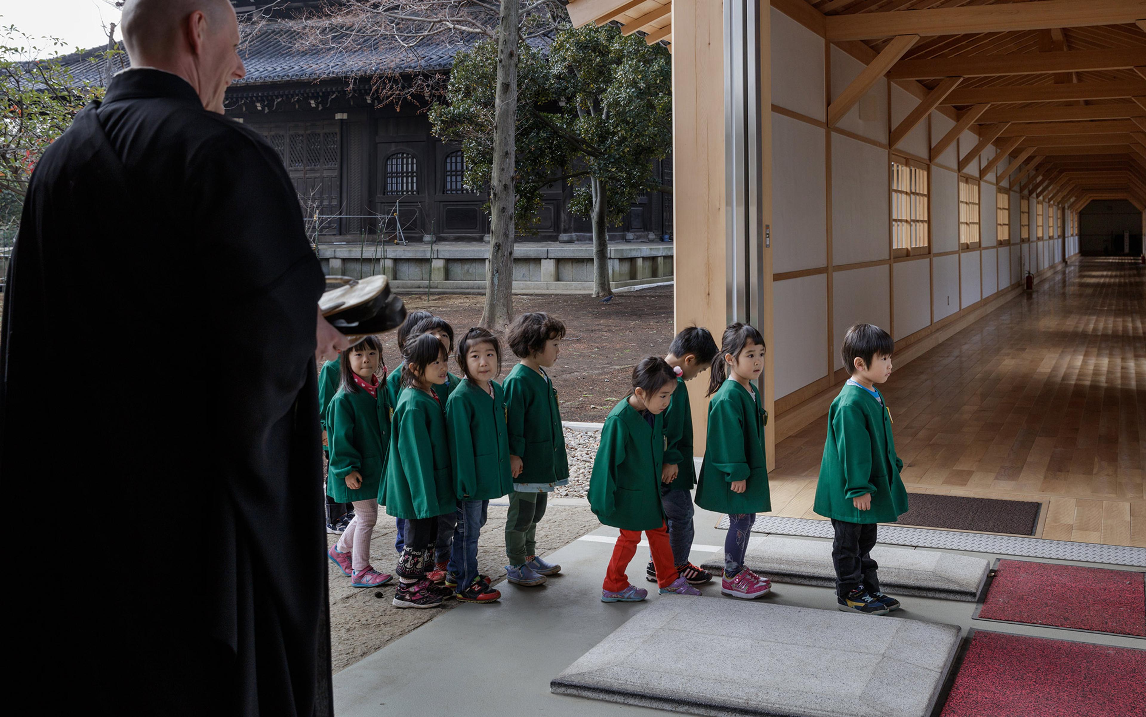 Young children wearing green jackets queuing in a line indoors supervised by a person in black robes, with a traditional building in the background.