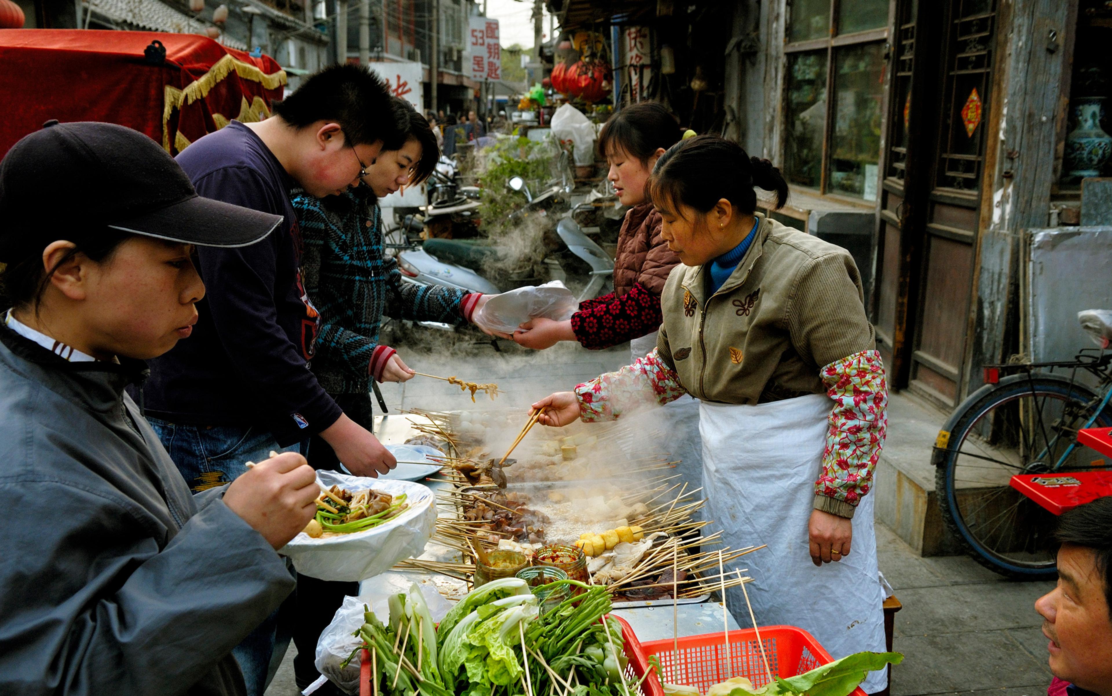 Street food vendor cooking skewers on a grill as people gather to eat and buy in a bustling outdoor setting.