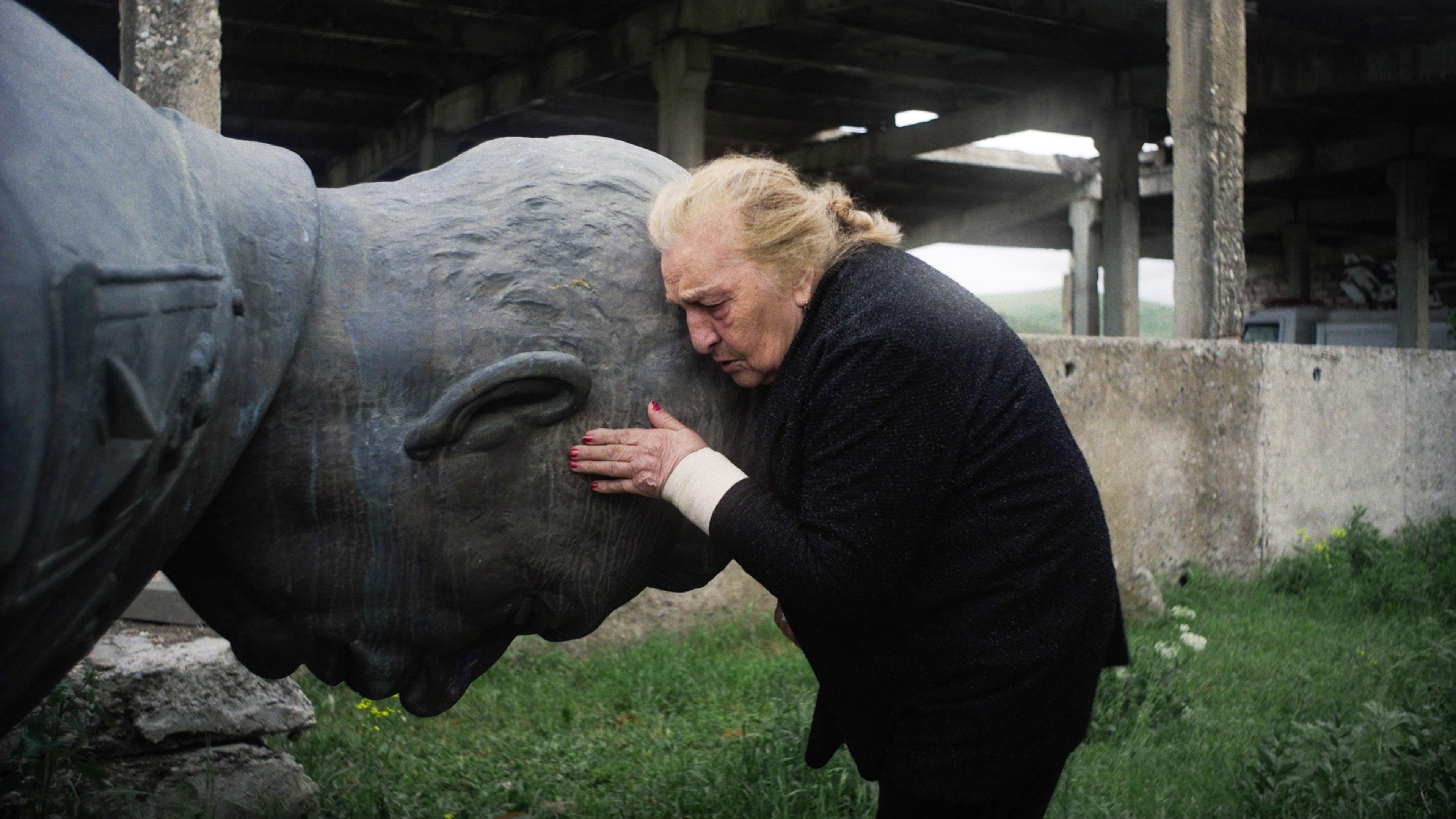 Photo of an elderly woman resting her forehead on a large stone bust outdoors near a decaying concrete structure.