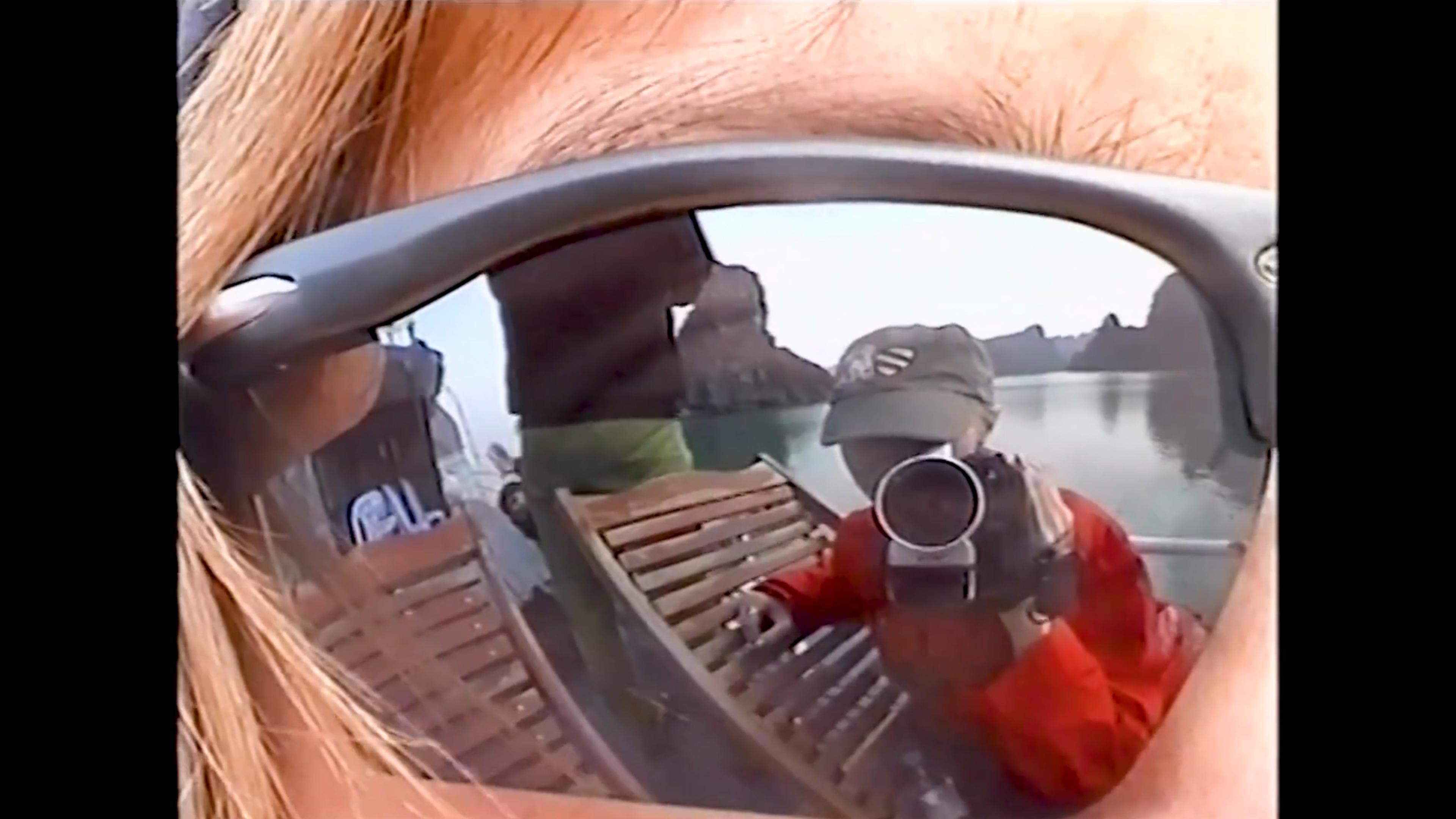 A video camera reflected in sunglasses with a scenic view of water and hills in the background.