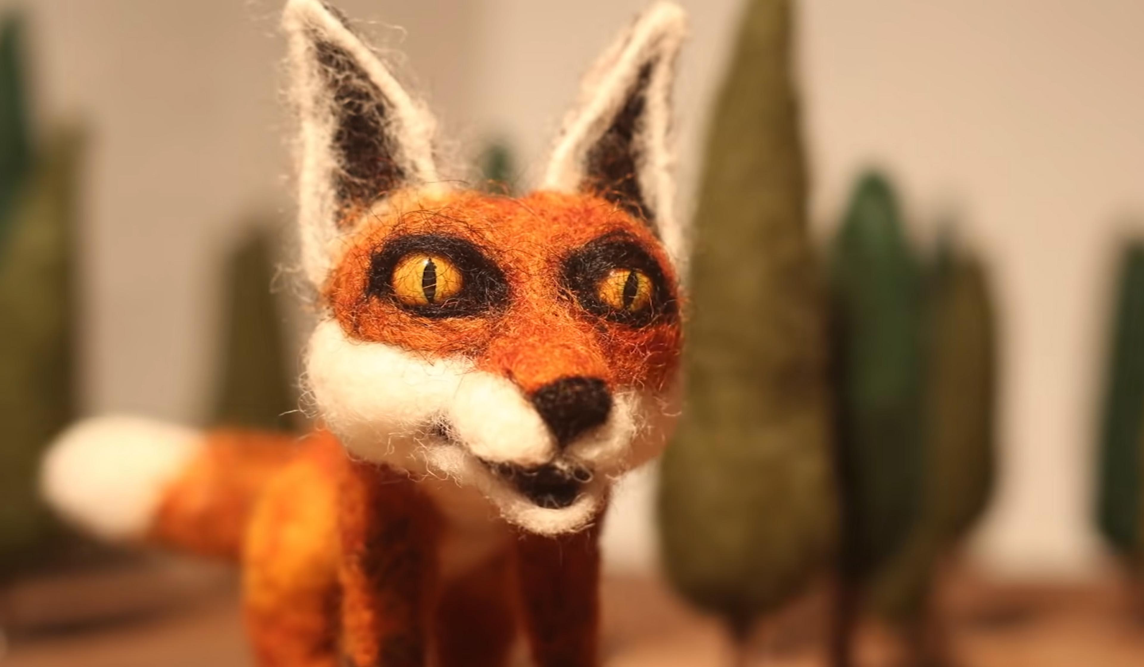 A felted wool fox sculpture with bright yellow eyes stands in front of a blurred background of green trees.