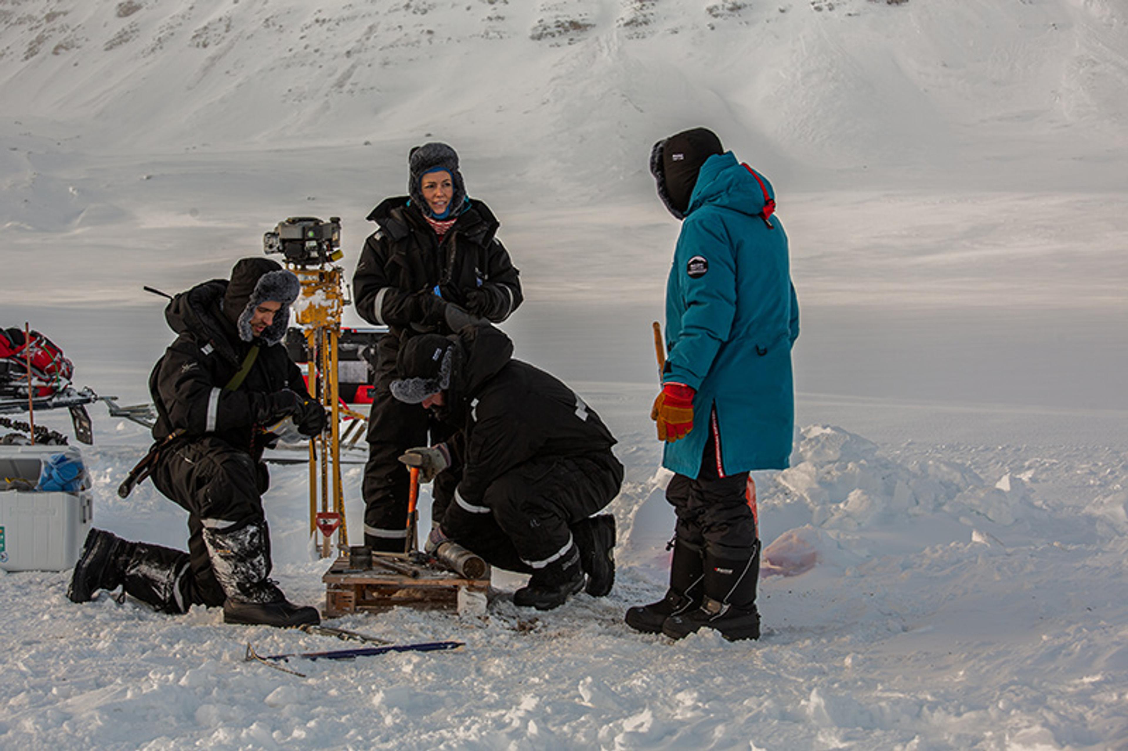 Photo of four people in winter gear conducting a task in snowy terrain, with equipment and a mountainous background.