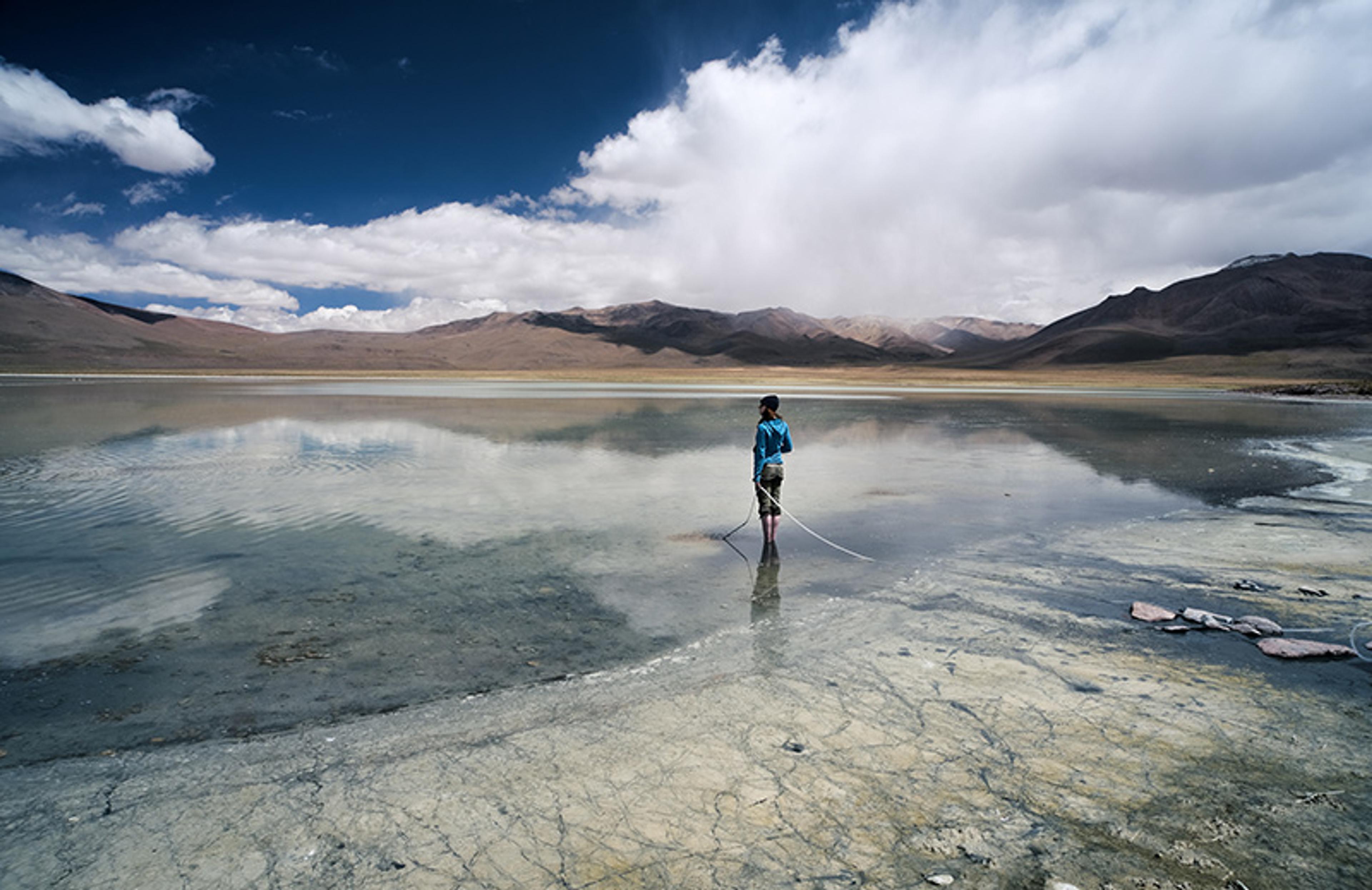Photo of a person standing on a reflective, shallow lake with distant mountains under a blue sky with clouds.