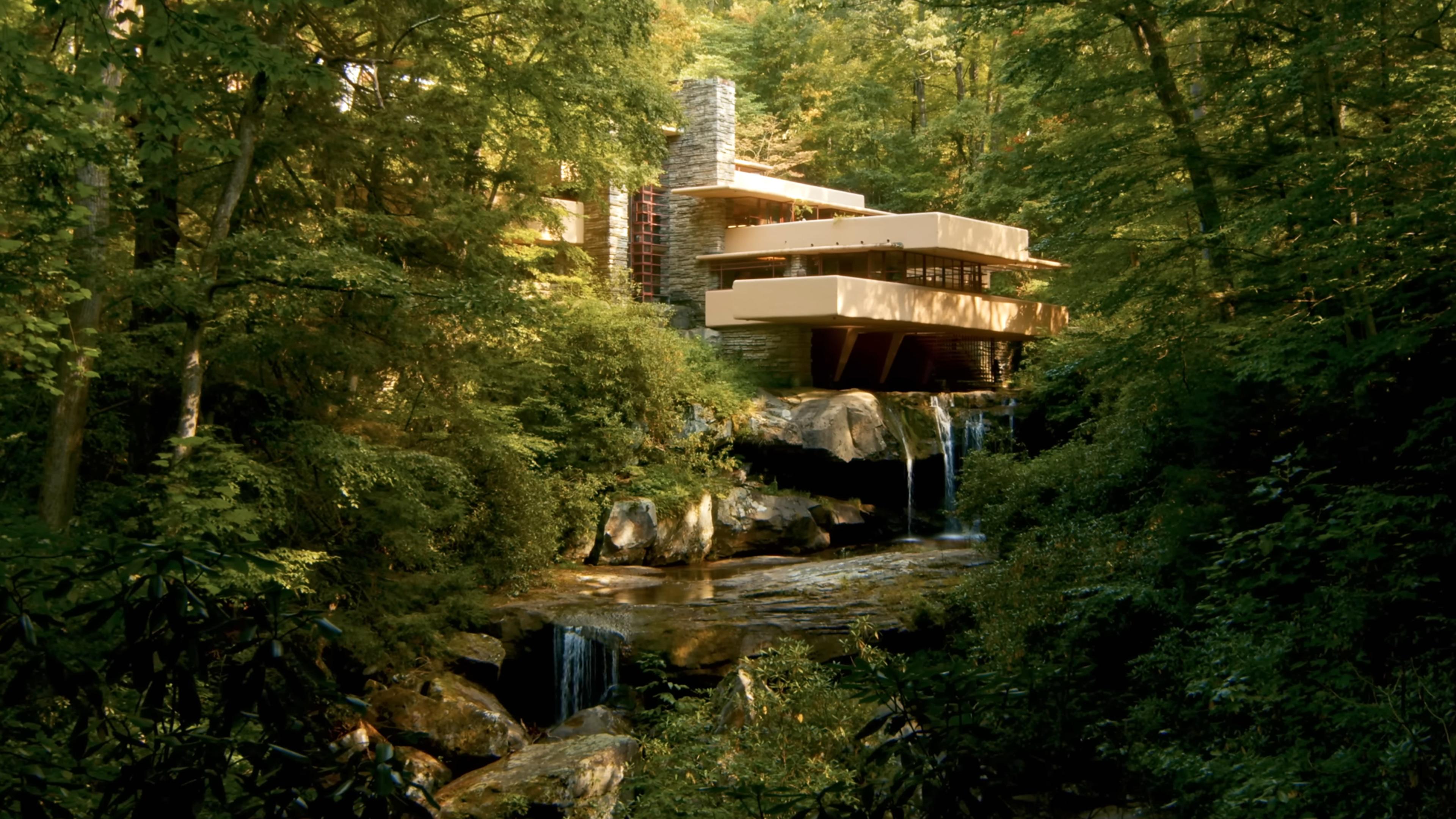 Photo of a modern house built over a waterfall, surrounded by dense green forest and a rocky stream.