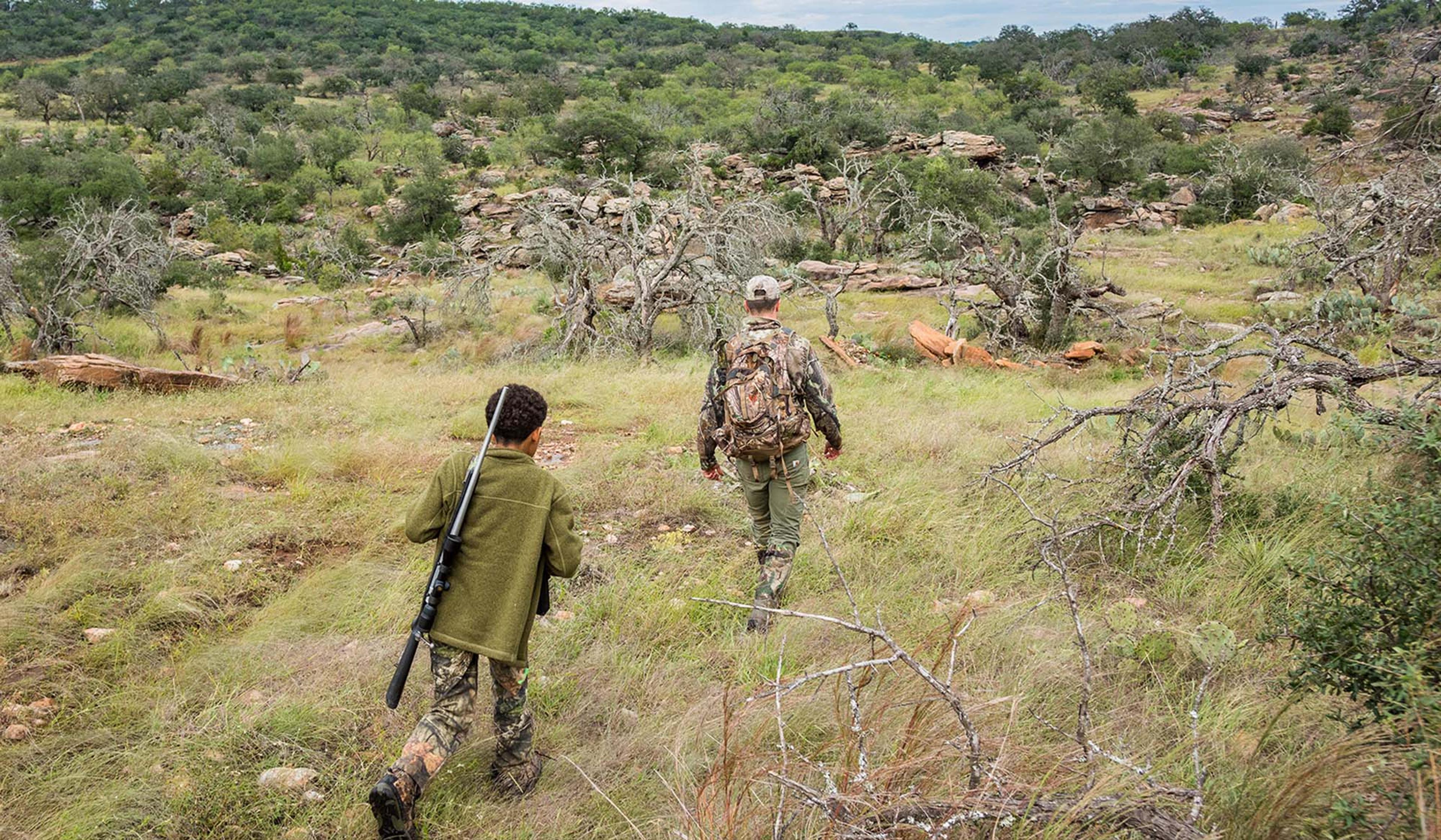Two hunters hiking through a grassy, rocky landscape with trees. One carries a rifle, the other a backpack.