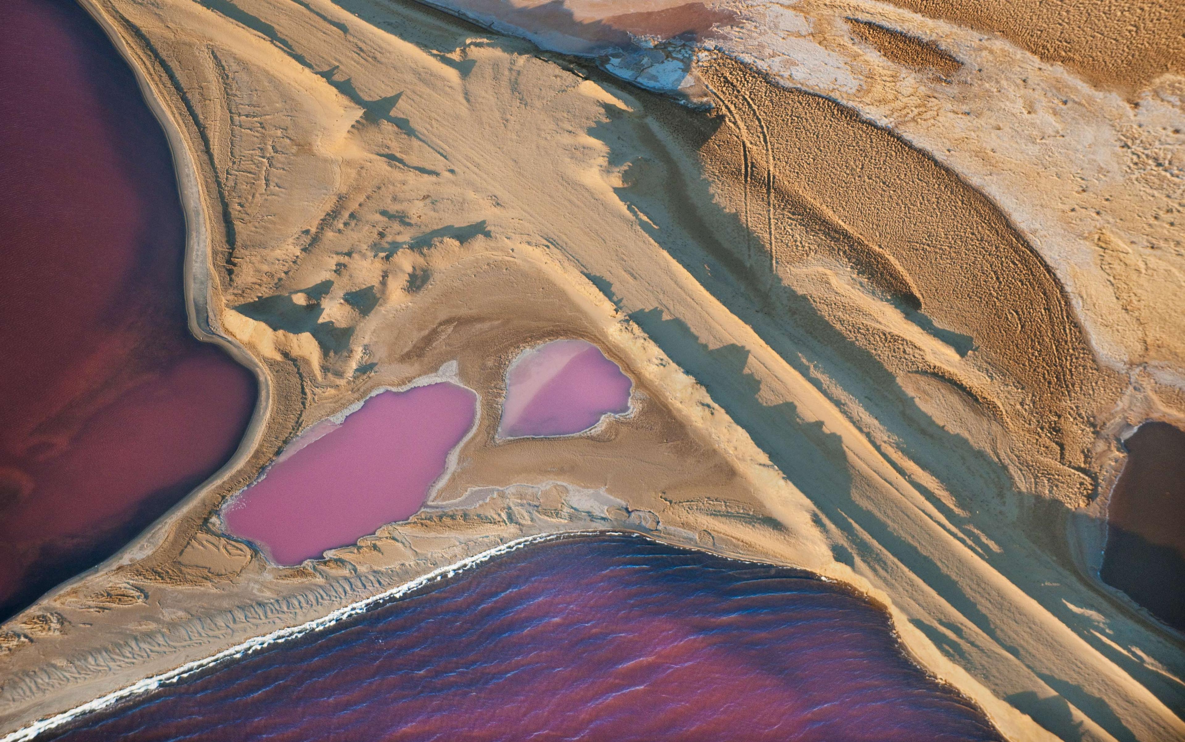 Aerial photo of a landscape with pink and purple water bodies amid sandy terrain. The contrast creates unique patterns and textures.