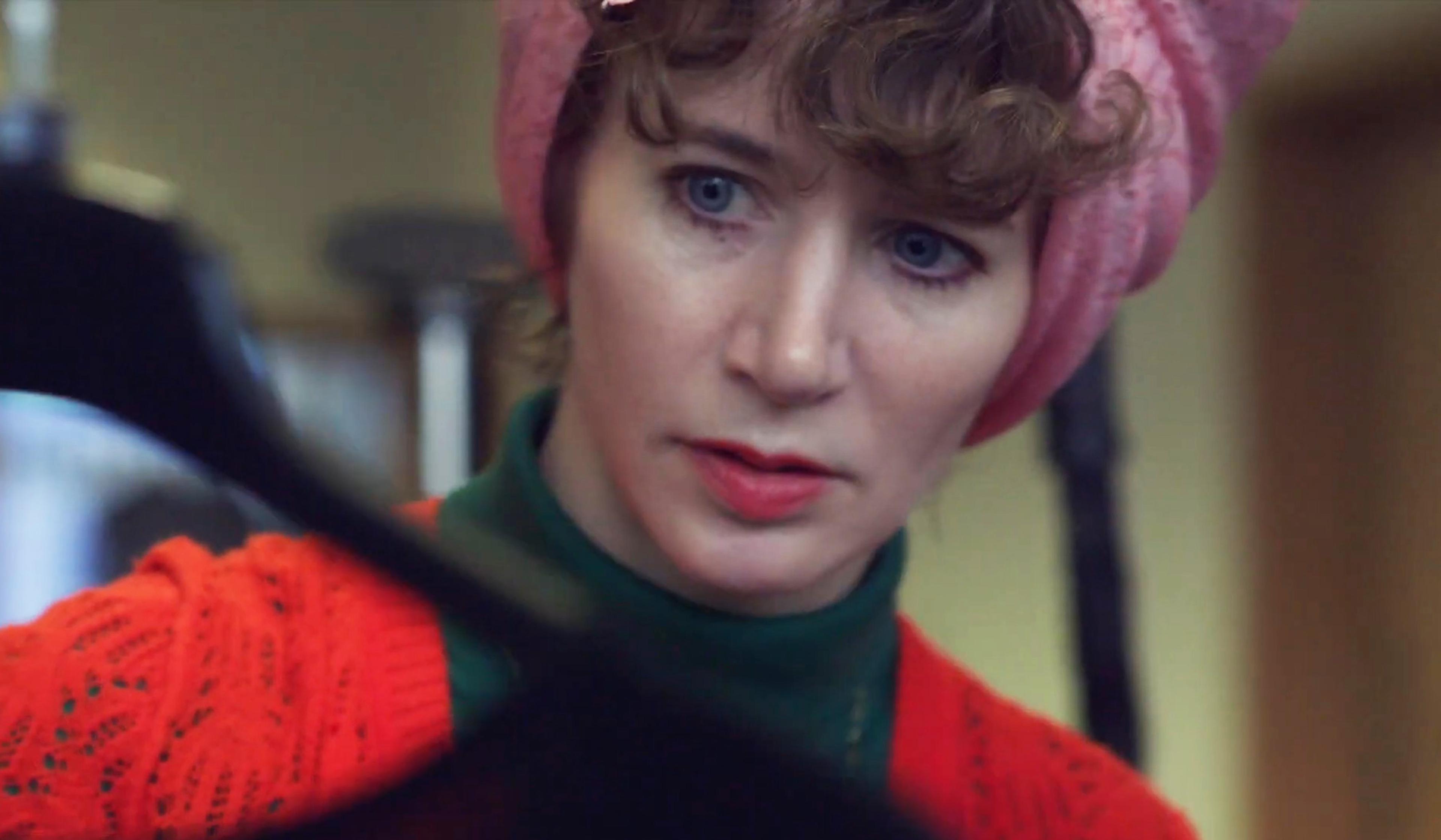A woman in a pink headscarf and red cardigan who is Miranda July looking intently at something off-camera.
