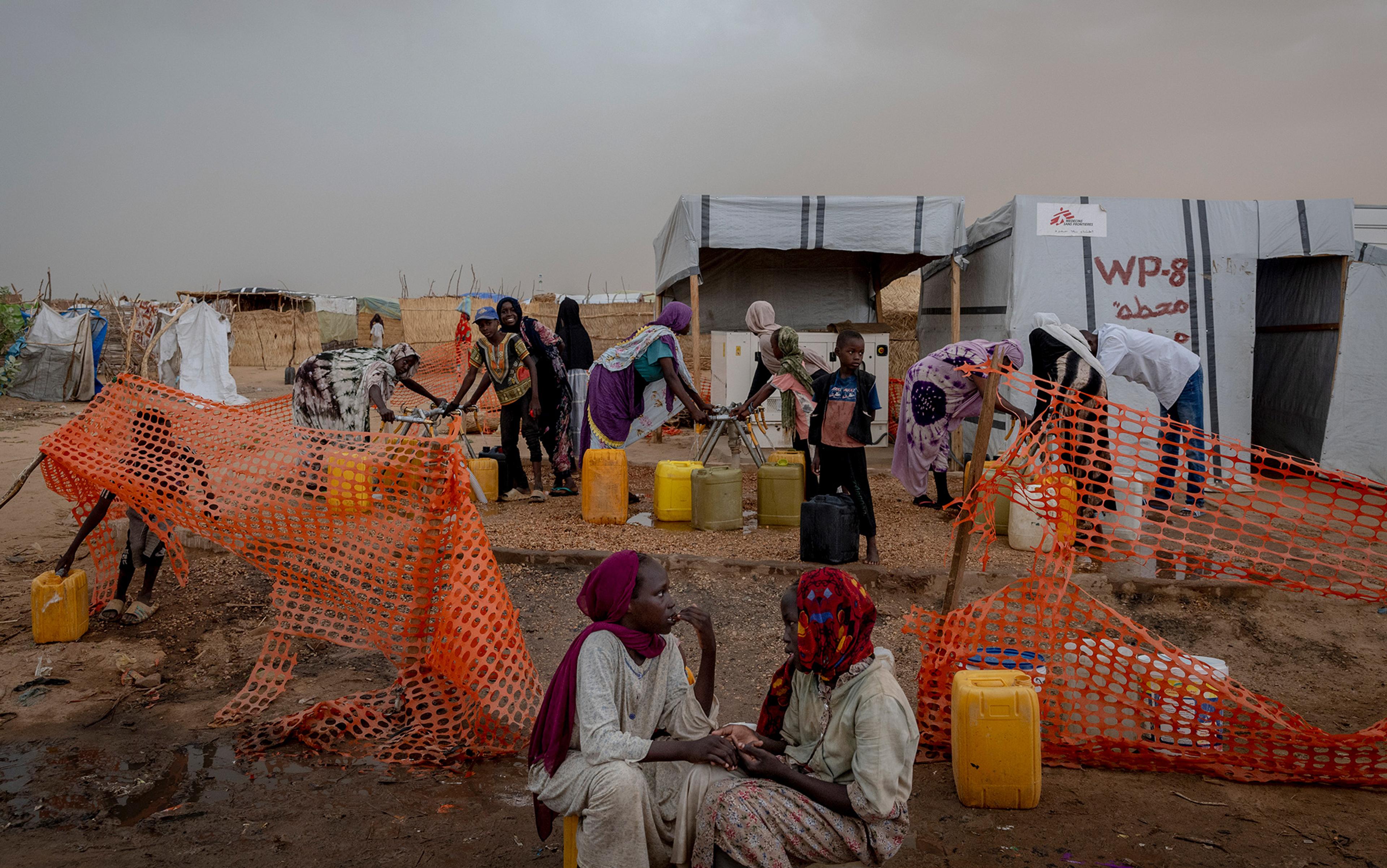 People collecting water from a makeshift station in a camp, surrounded by orange netting and tents in the background.
