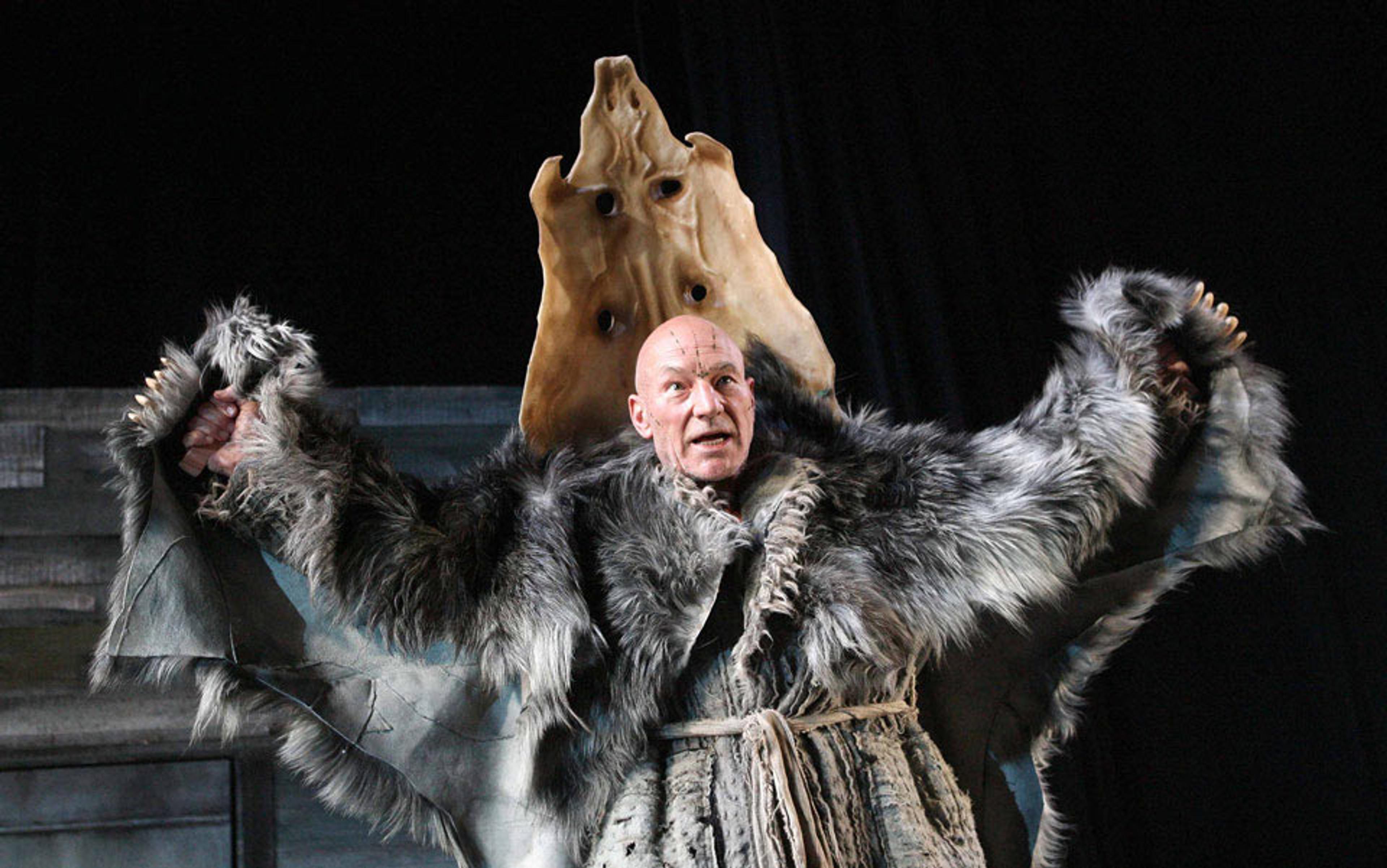 An actor on stage wearing a fur robe and a mask on his back, with arms raised in an expressive pose.