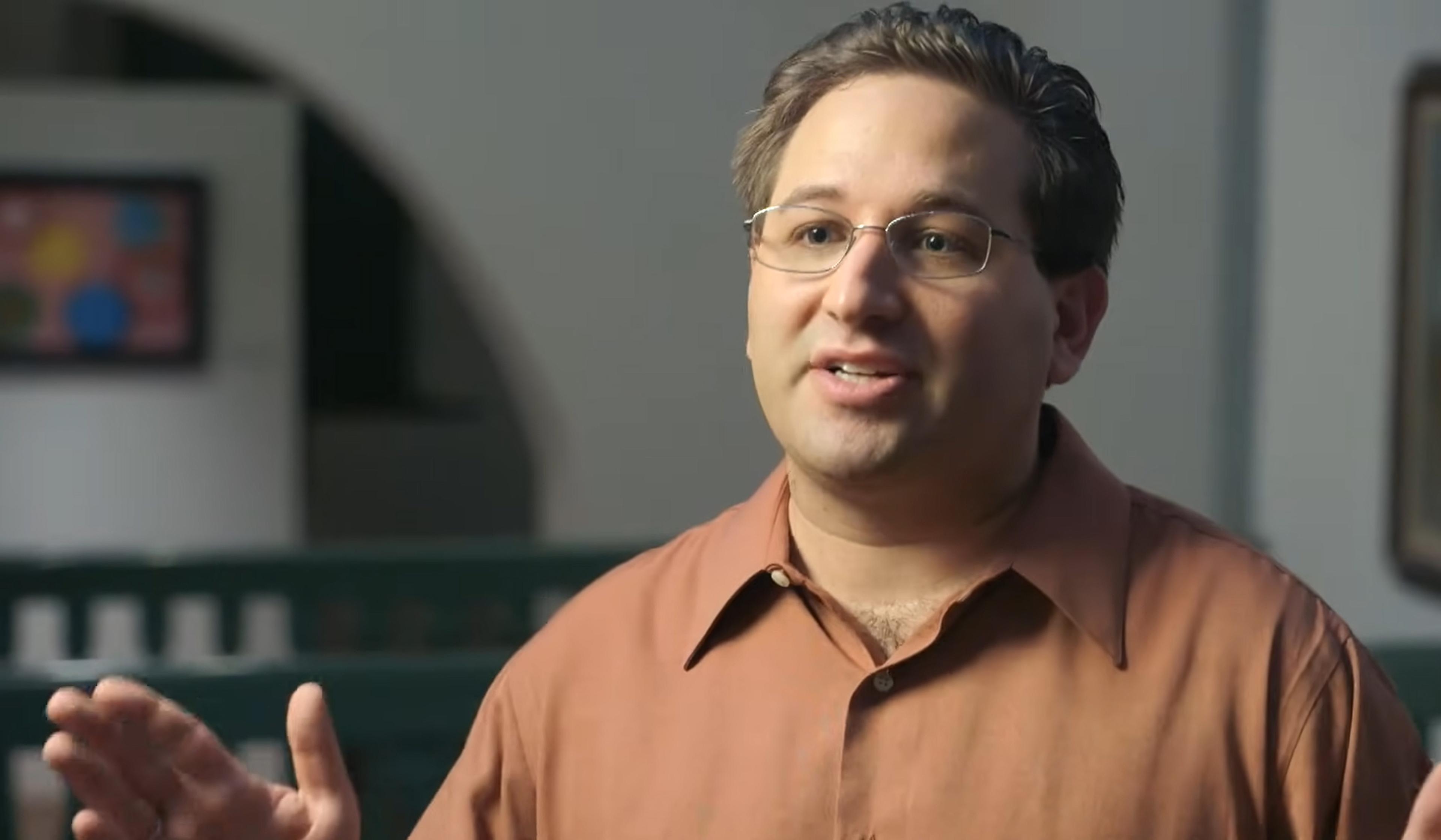 Man with glasses speaking, wearing a brown shirt. Background is blurred with a colourful painting visible.